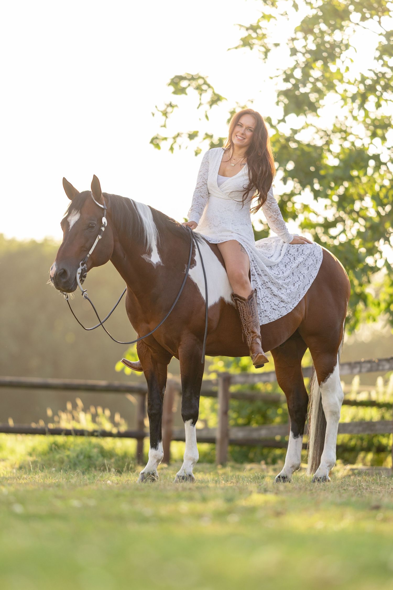 A smiling woman in a white dress rides a brown and white paint horse in a sunny outdoor paddock.