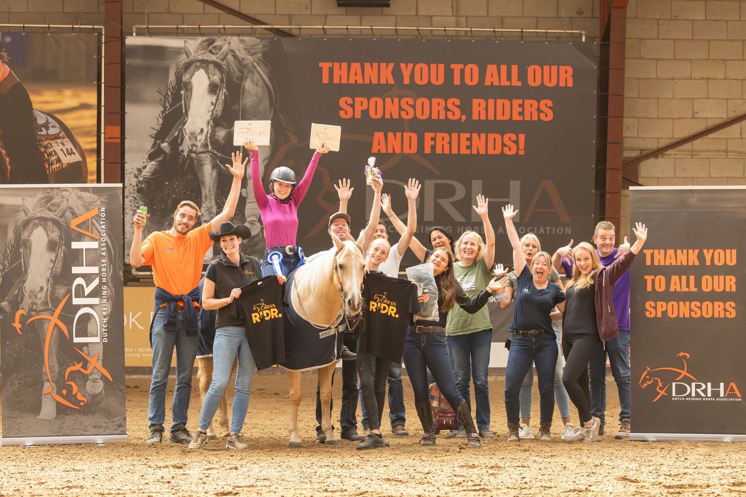 Large group celebrating with a horse at an equestrian event in front of a sponsor wall.