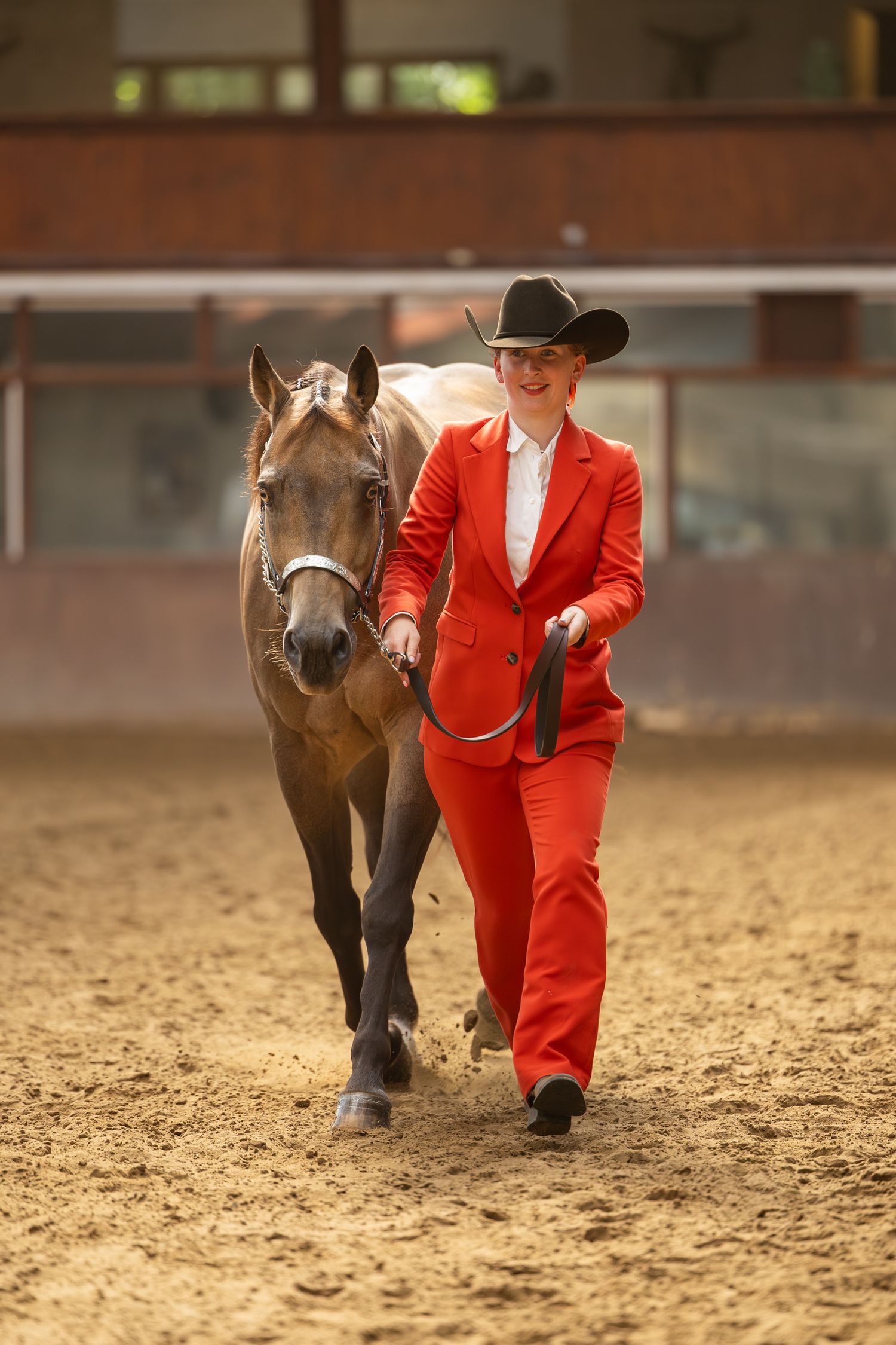 Handler in bright red suit leading bay horse through indoor arena presentation.