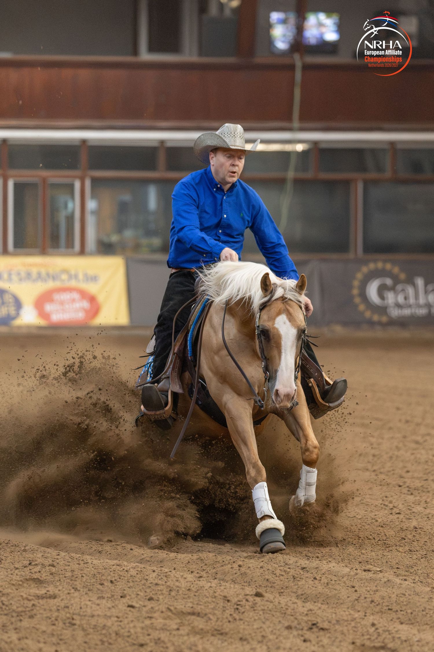 Cowboy in blue shirt riding palomino horse in NRHA reining competition on dirt arena.