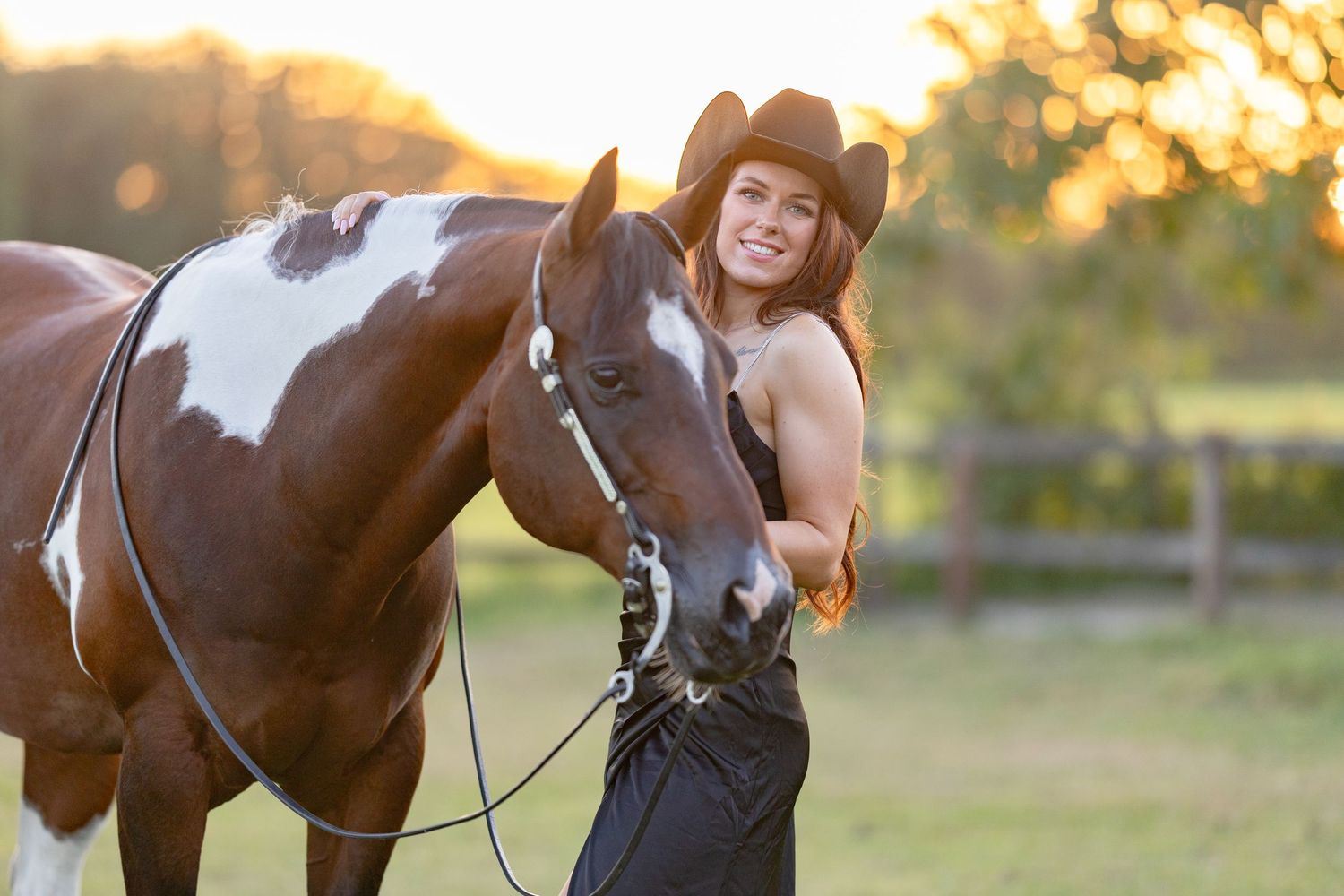 Young woman in cowboy hat smiling beside a paint horse at golden hour on a ranch.