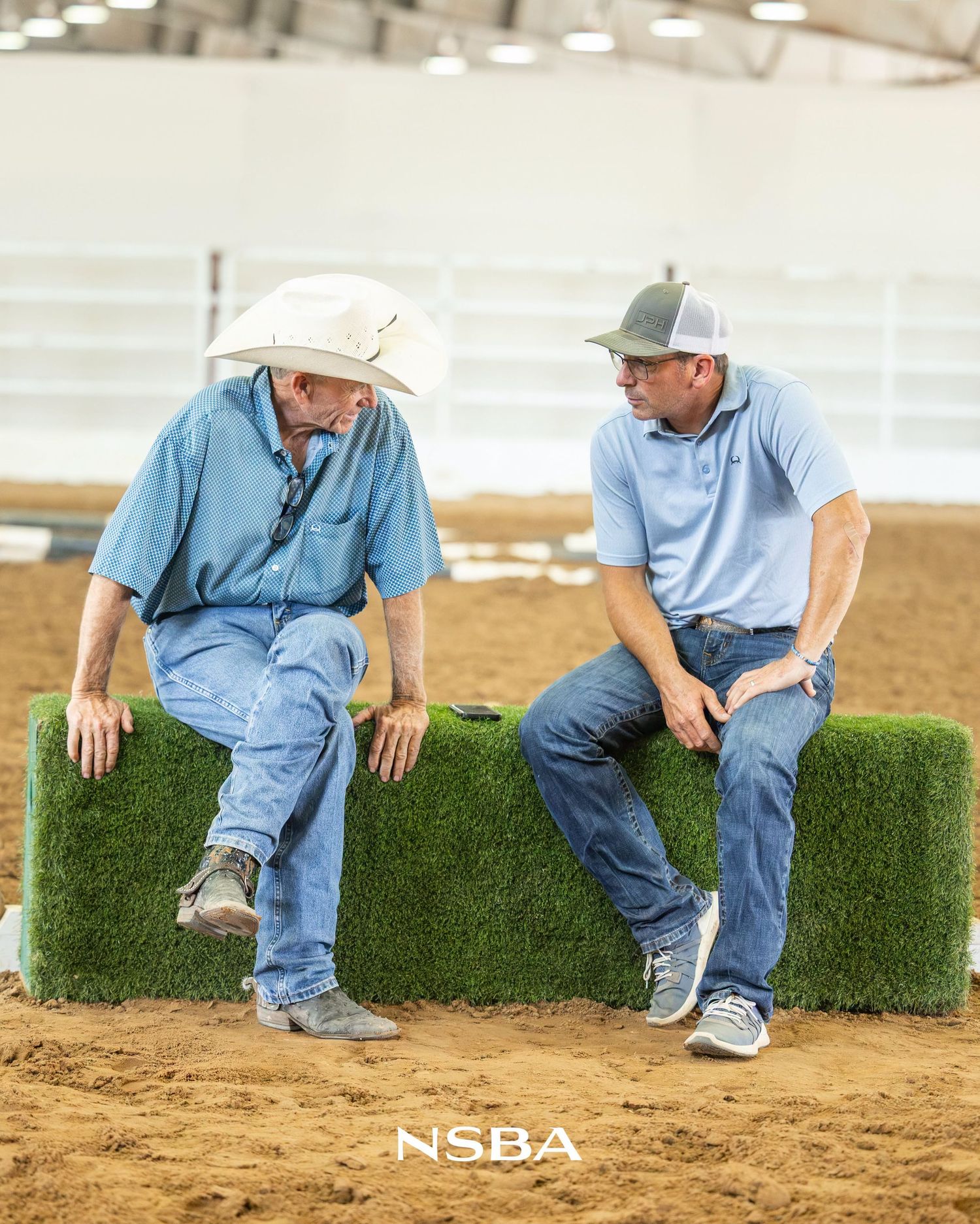Two men in blue shirts and jeans sit on hay bales chatting at an NSBA horse show arena.