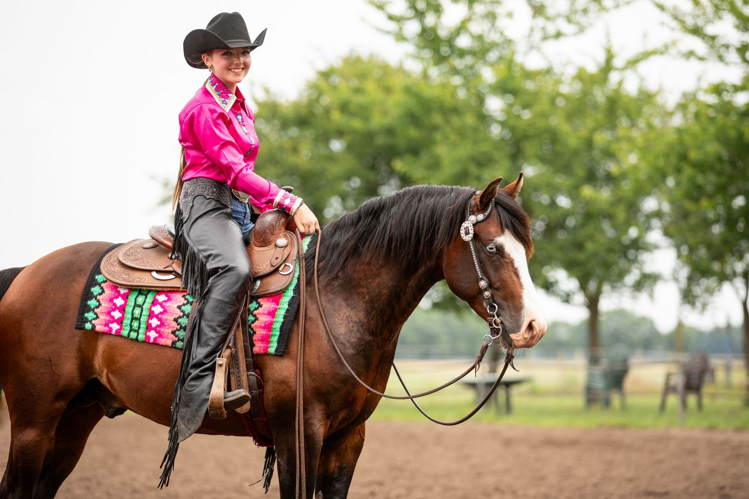 Rider in pink western attire sits atop a dark horse with decorative saddle blanket in outdoor arena.