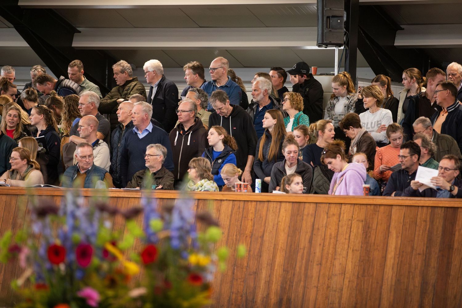 A crowd of spectators watches intently from wooden bleachers with colorful flowers in the foreground.