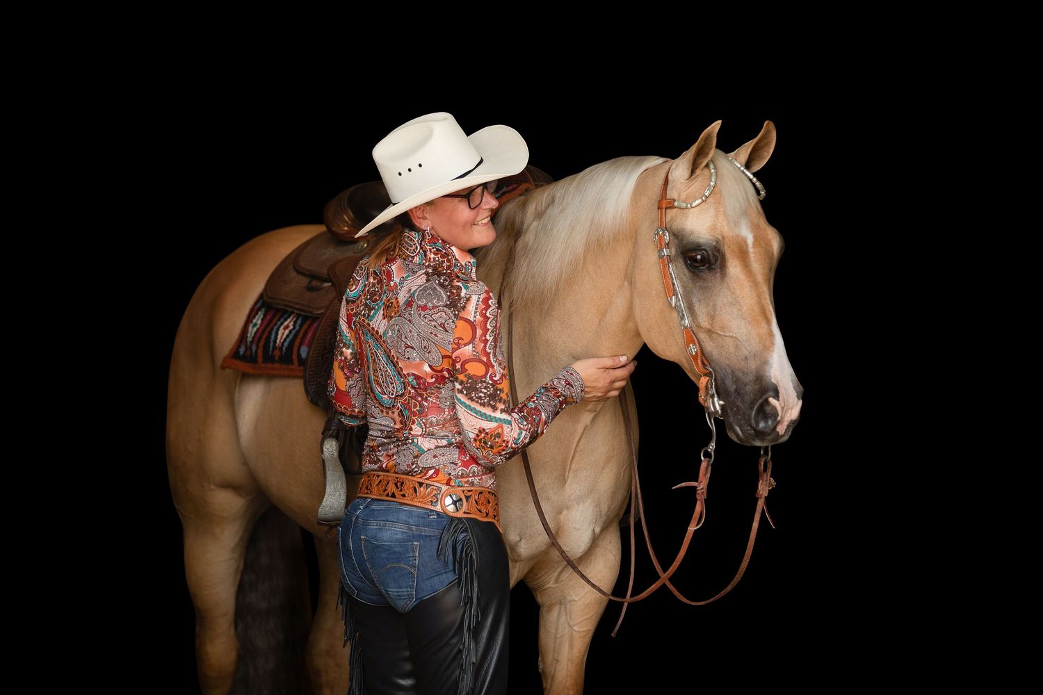 Cowboy in white hat and floral shirt stands beside a palomino horse against a black background.