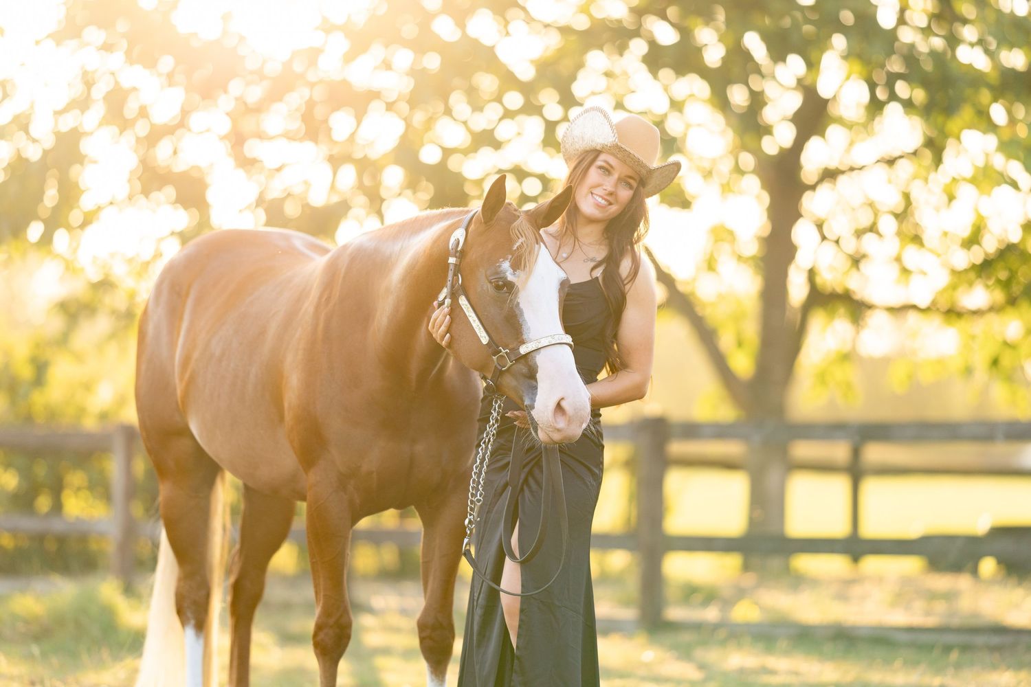 A young woman in a cowboy hat and black dress stands beside a brown horse in golden sunlight near a wooden fence.