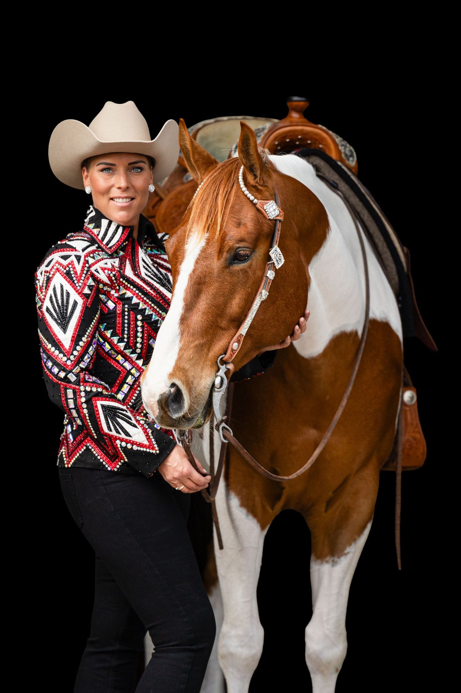 Woman in patterned western shirt and cowboy hat poses with a chestnut and white Paint horse with decorative bridle.