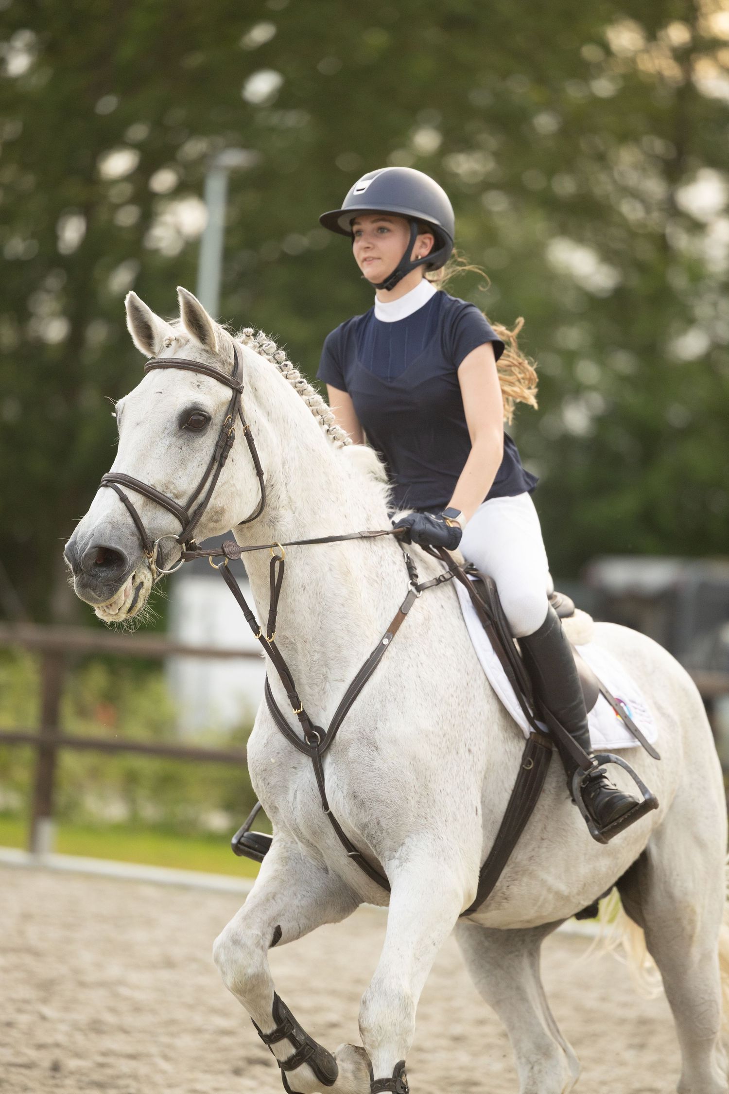Young woman in equestrian helmet and white breeches riding a white horse in an outdoor arena.