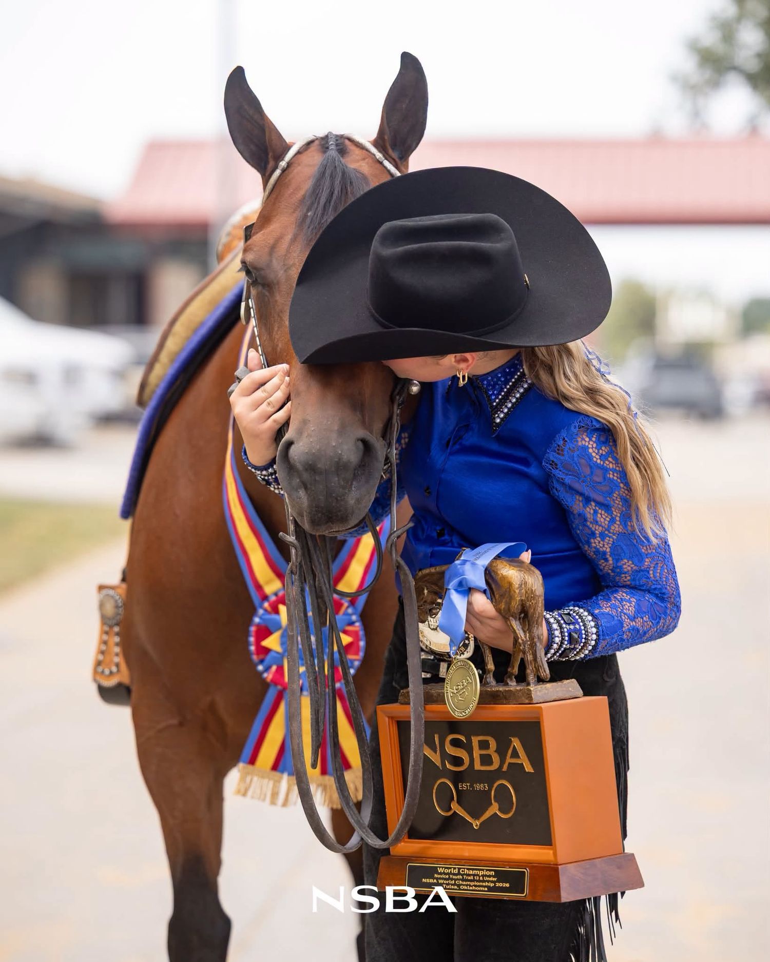 A woman in a blue lace top and black cowboy hat embraces her horse beside an NSBA trophy with colorful ribbons.