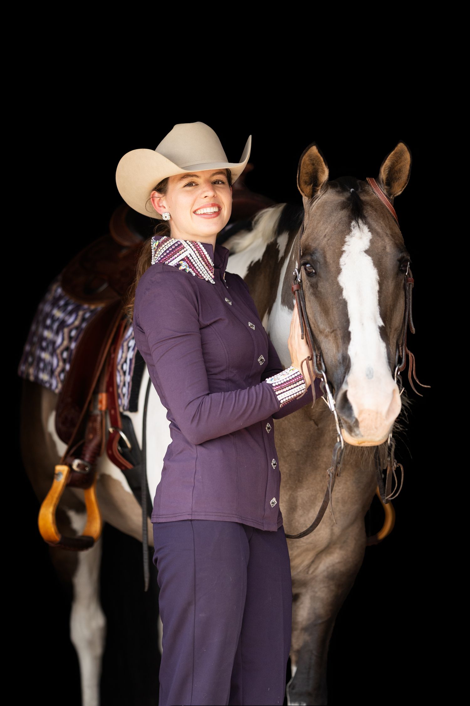 A paint horse stands with a rider in a purple show outfit against a black backdrop.