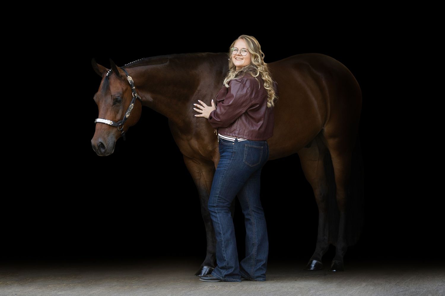 A blonde woman in a burgundy jacket stands beside a chestnut horse on a dark background.