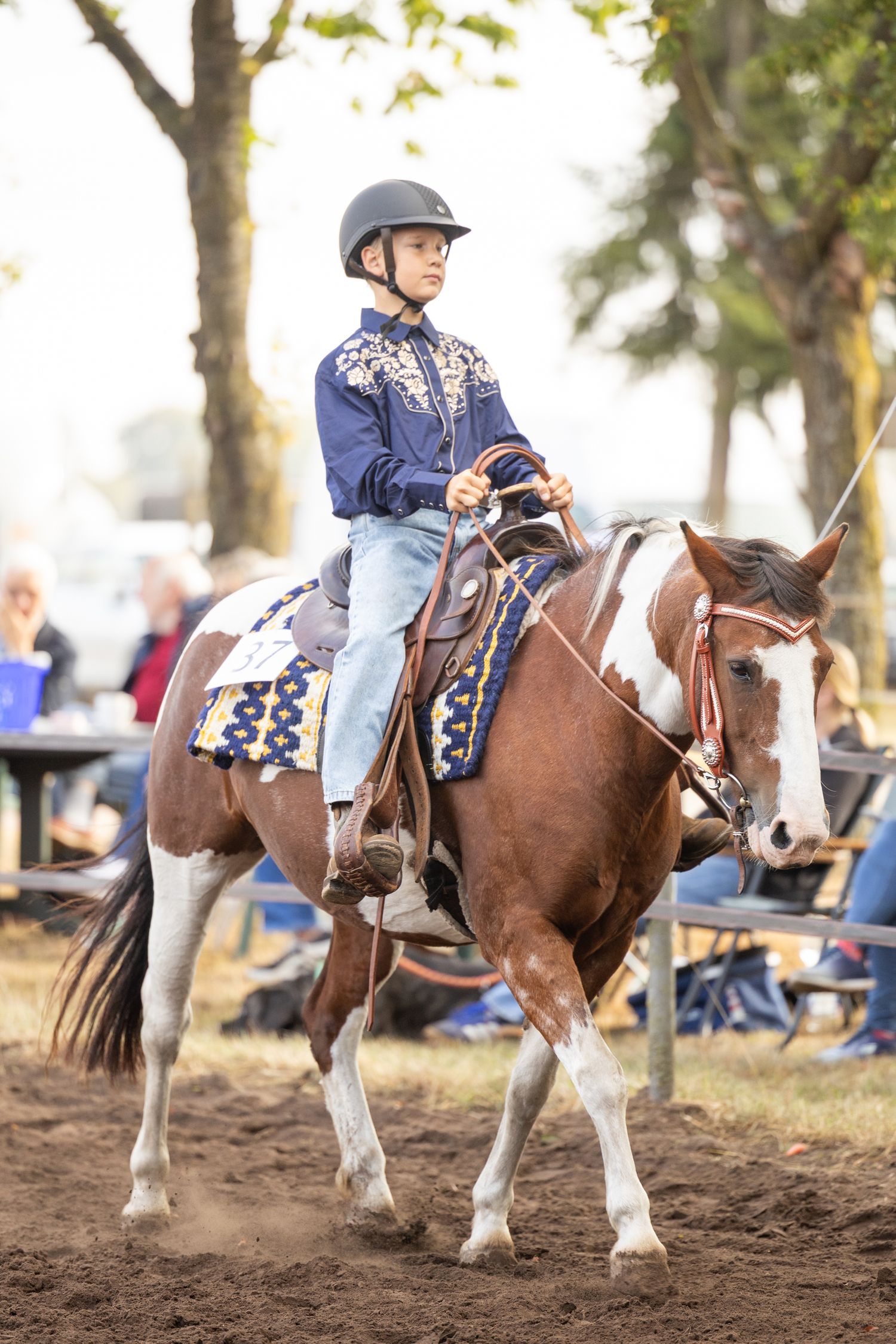 Equestrian rider in blue show outfit and helmet riding a paint horse with decorative western tack in an arena.