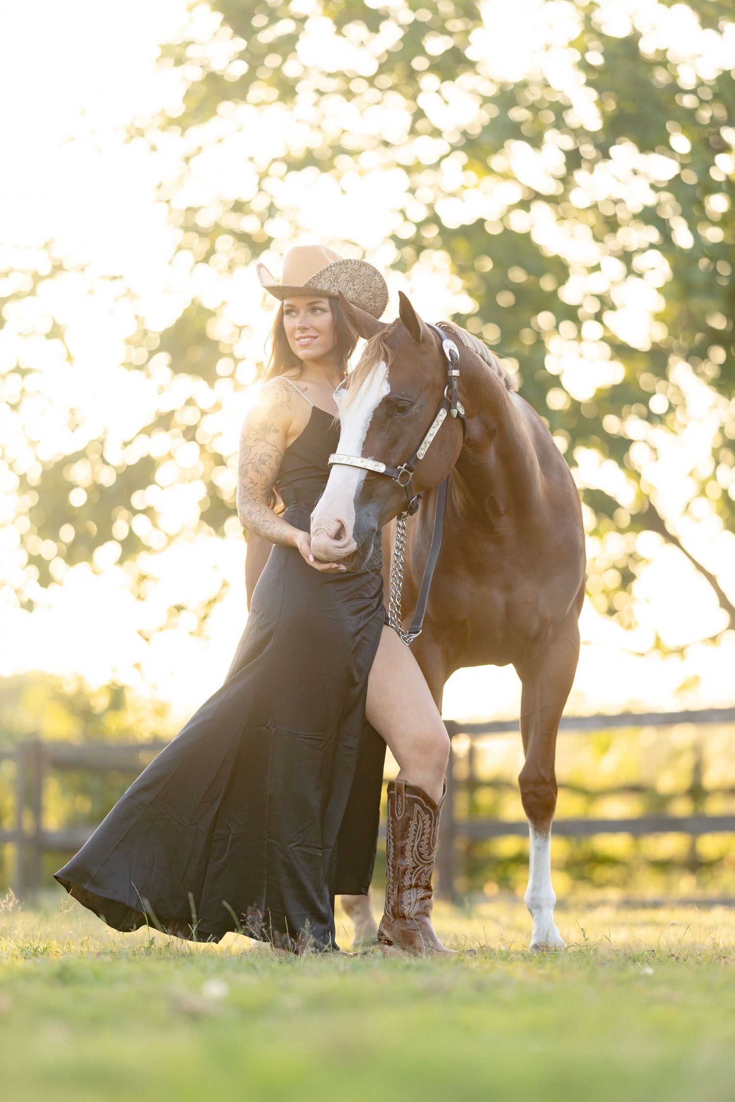 Woman in black dress and cowboy hat stands beside a brown horse in a sunlit outdoor setting.