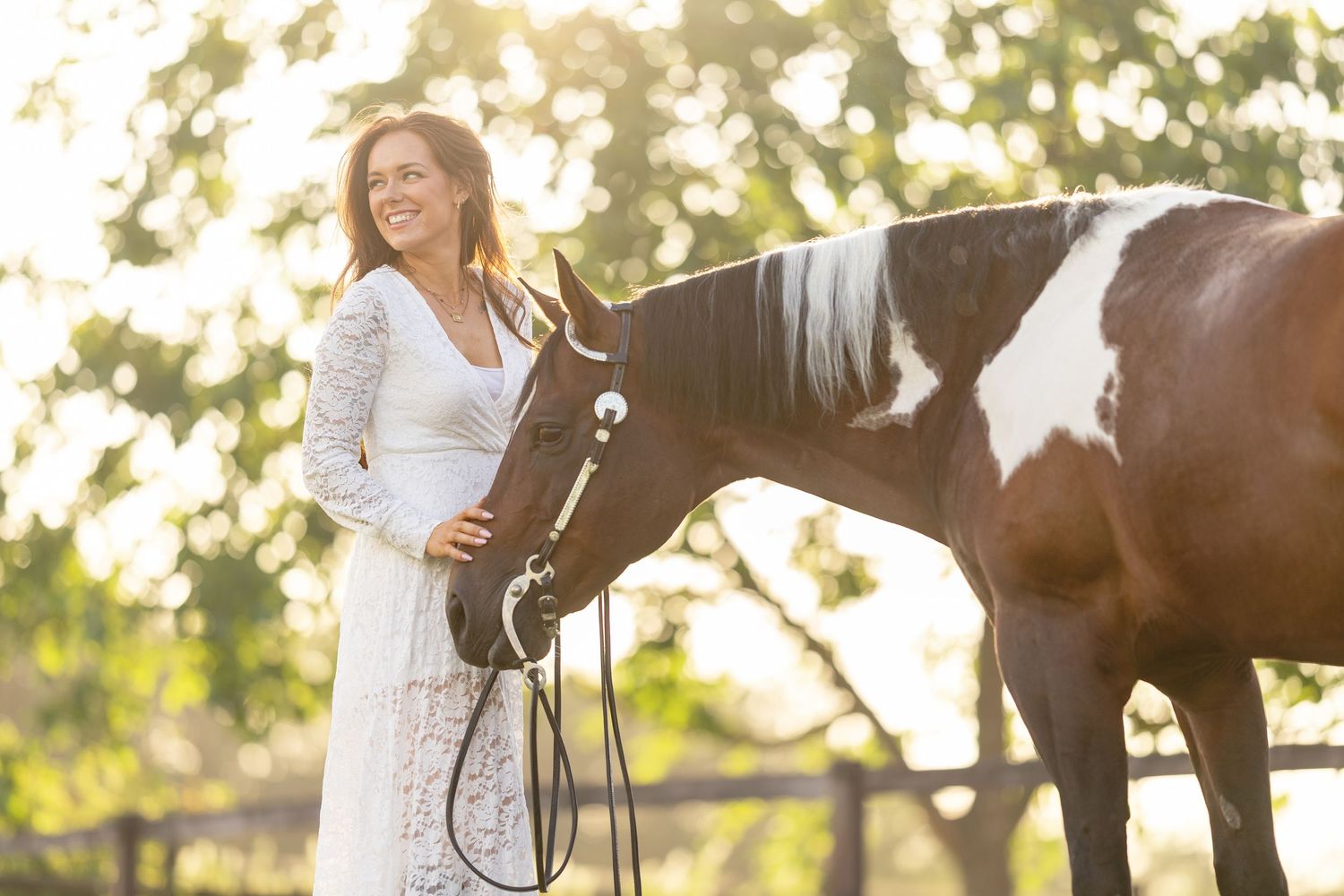 Woman in white lace dress smiling beside a paint horse outdoors in golden sunlight.