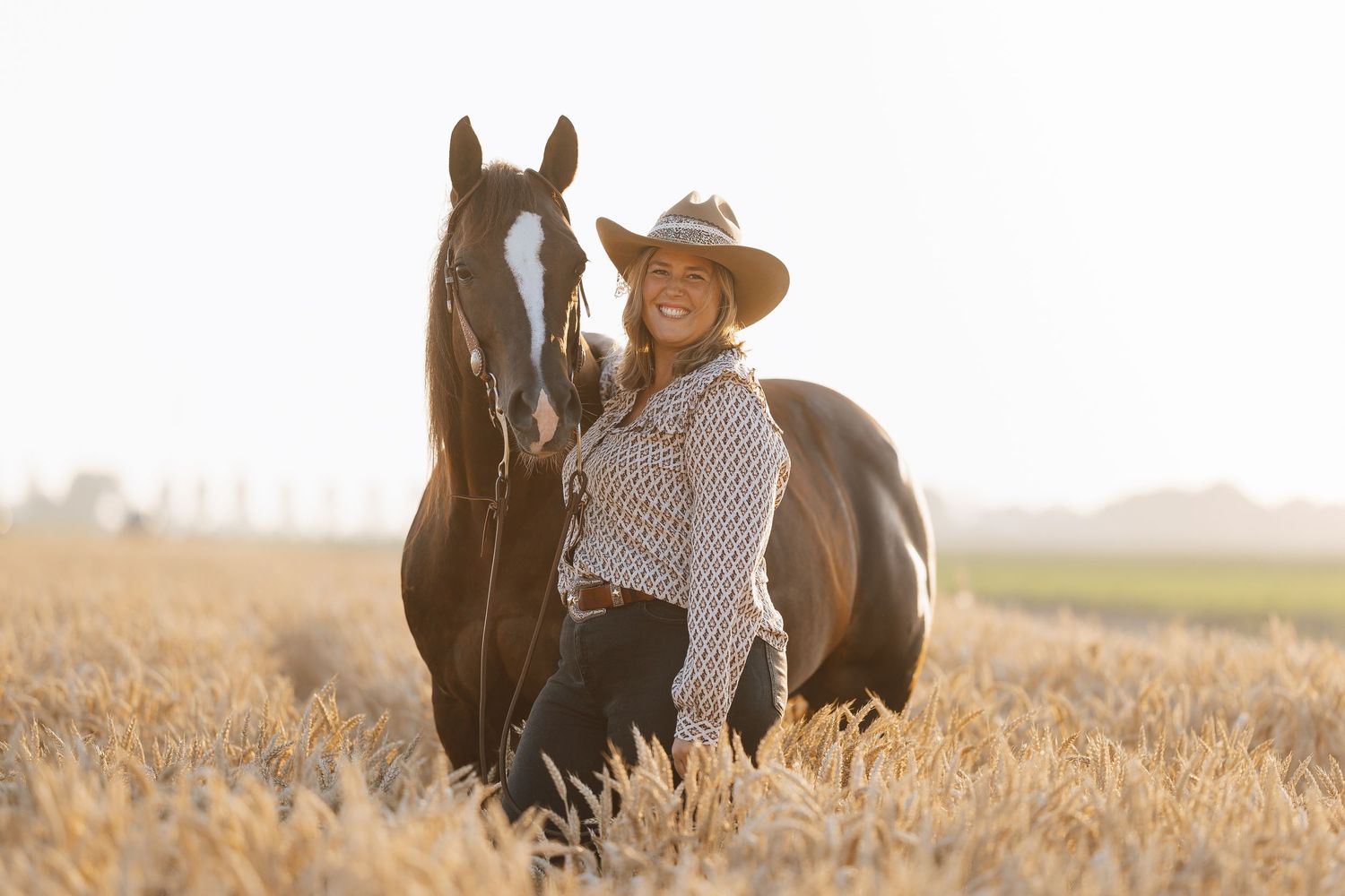 Smiling woman in cowboy hat standing beside a dark horse in a golden wheat field on a sunny day.
