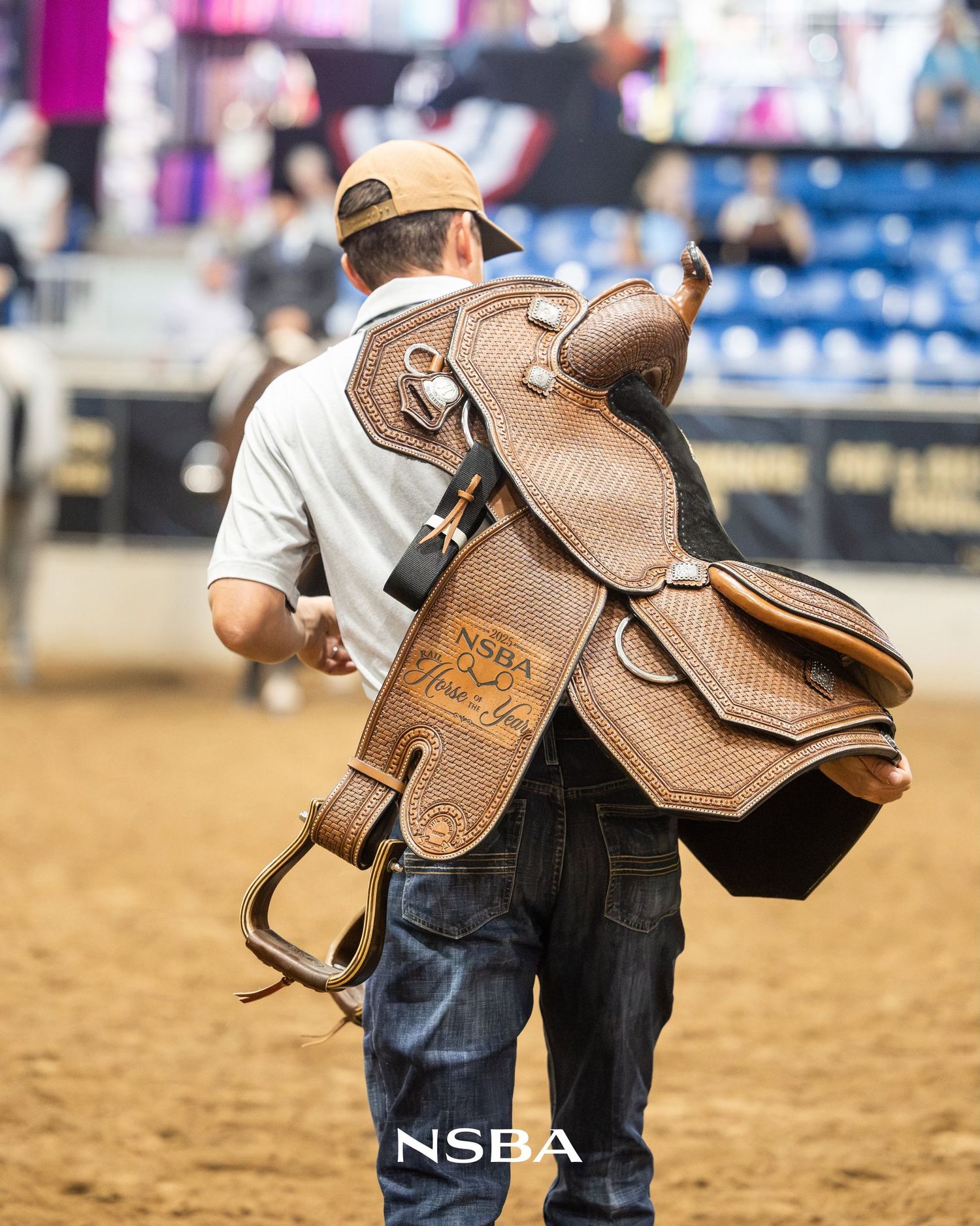 Man carrying an NSBA branded Western saddle at a rodeo arena competition.