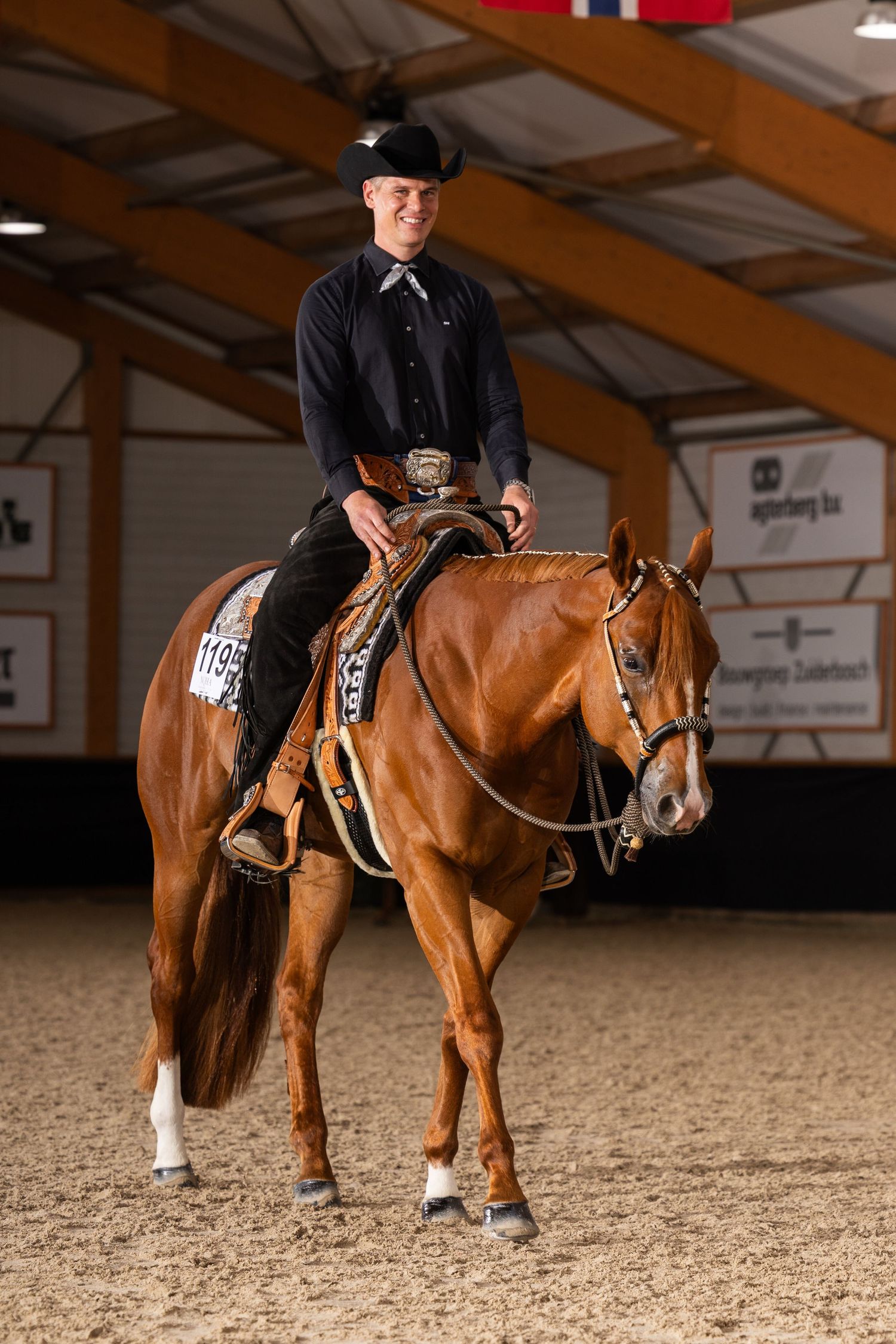 Equestrian rider in black attire atop a chestnut horse in an indoor arena during a competition.