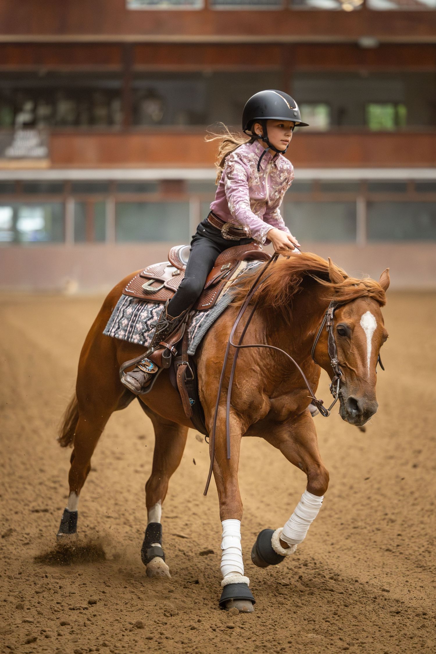 Young girl in helmet rides chestnut horse in indoor arena, competing in western equestrian event.