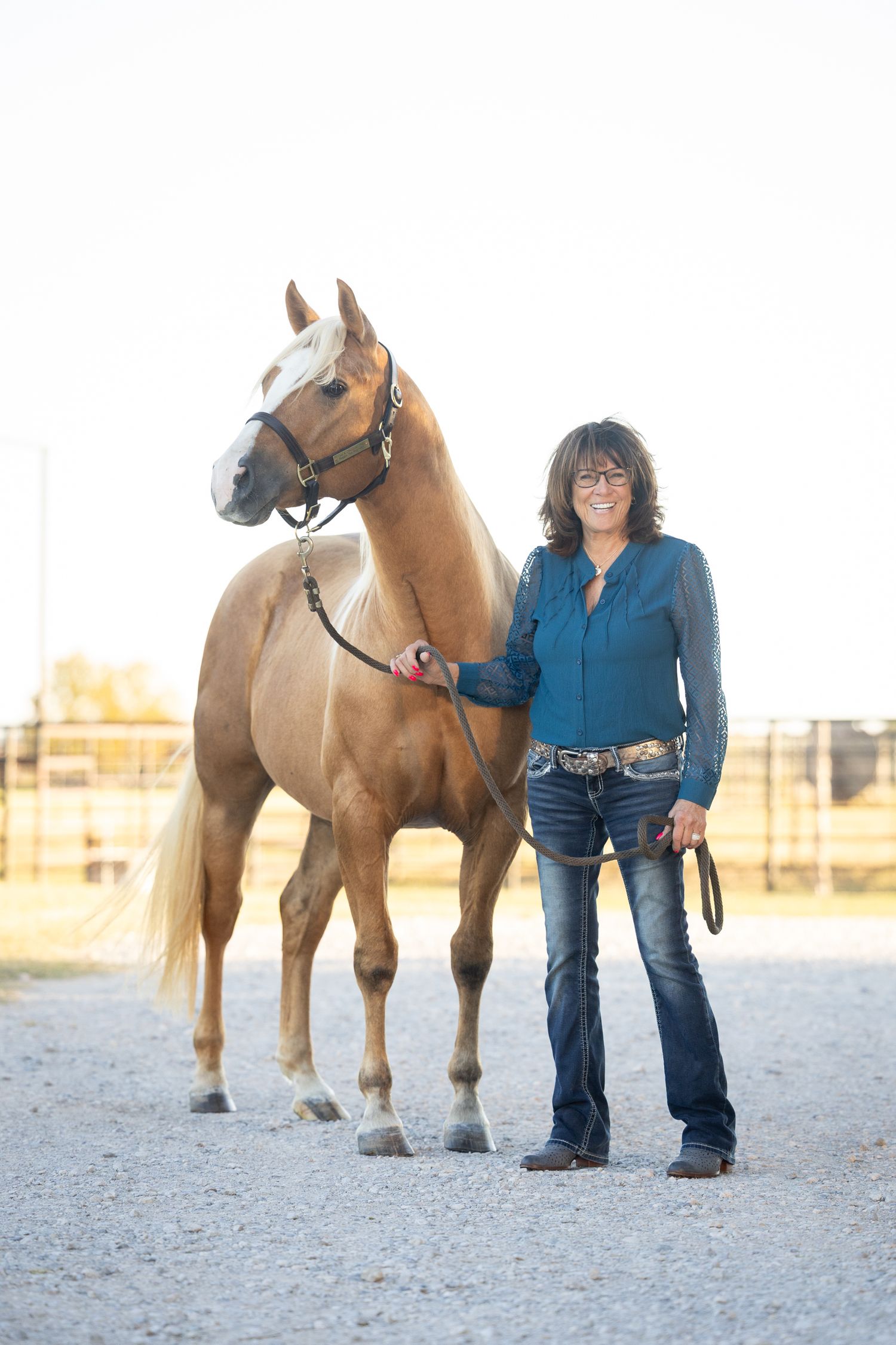 Person in blue shirt and jeans posing with brown horse in outdoor arena.