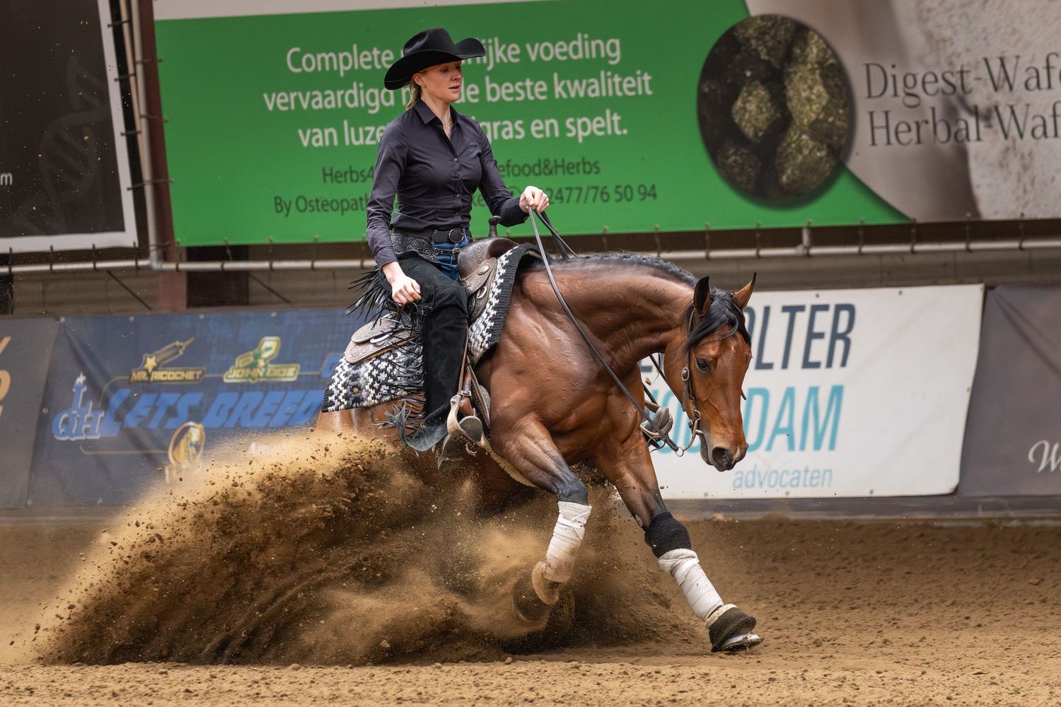 A rider guides their horse through a competitive barrel racing event in an indoor arena, kicking up dirt and dust.
