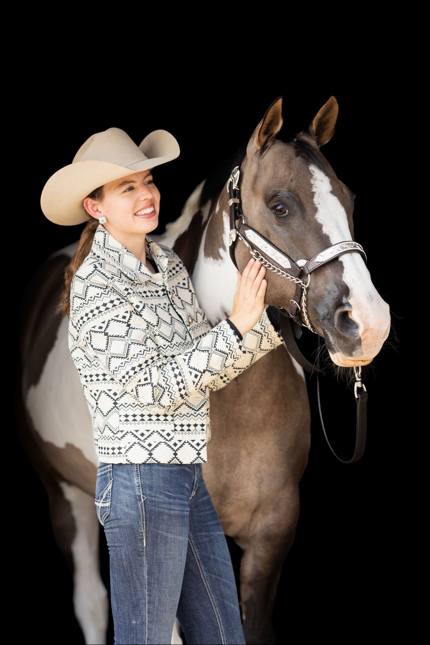 A paint horse poses with a rider wearing a patterned sweater and cowboy hat.