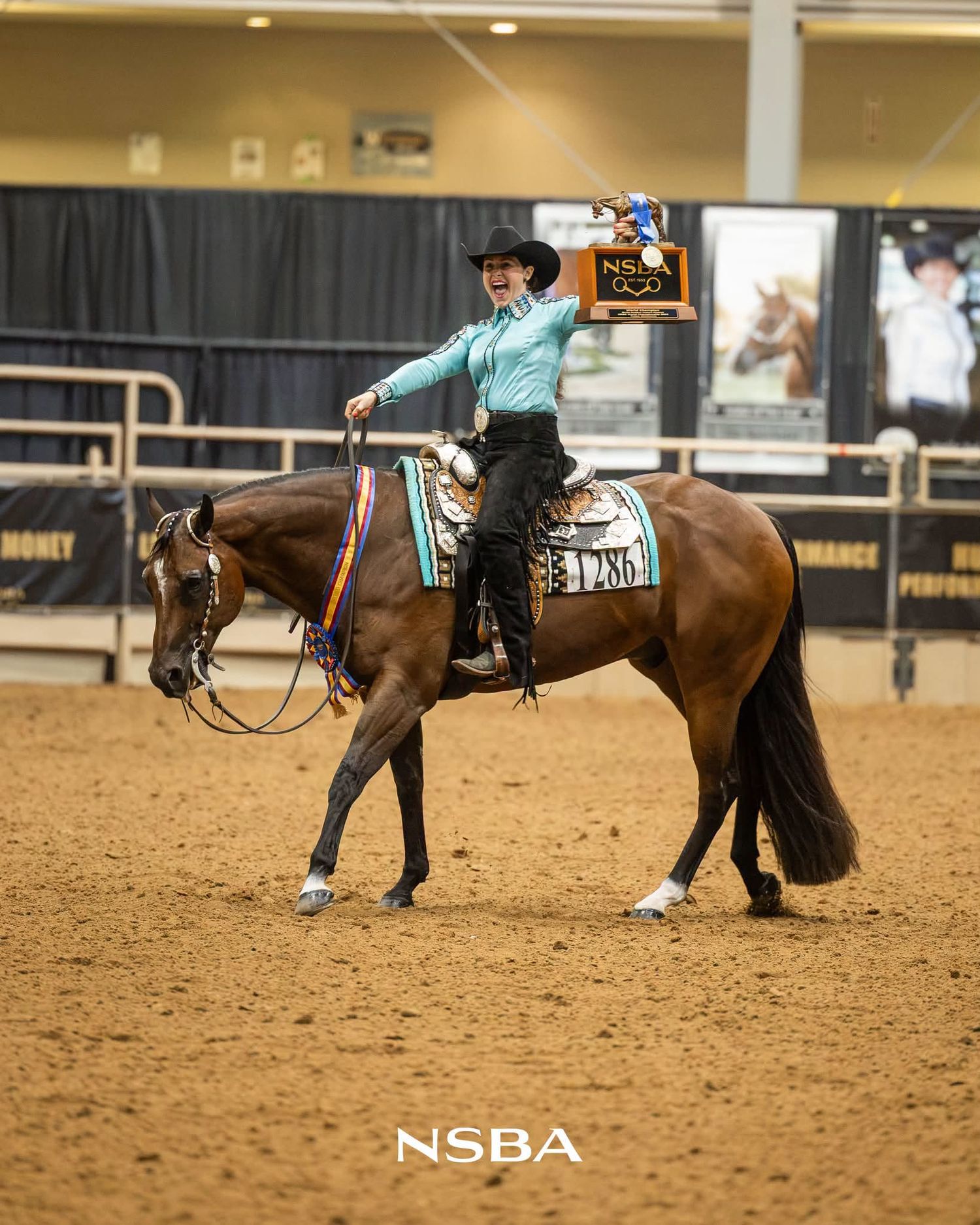 Rider in turquoise shirt holds trophy aloft on bay horse at NSBA equestrian competition on indoor arena dirt.