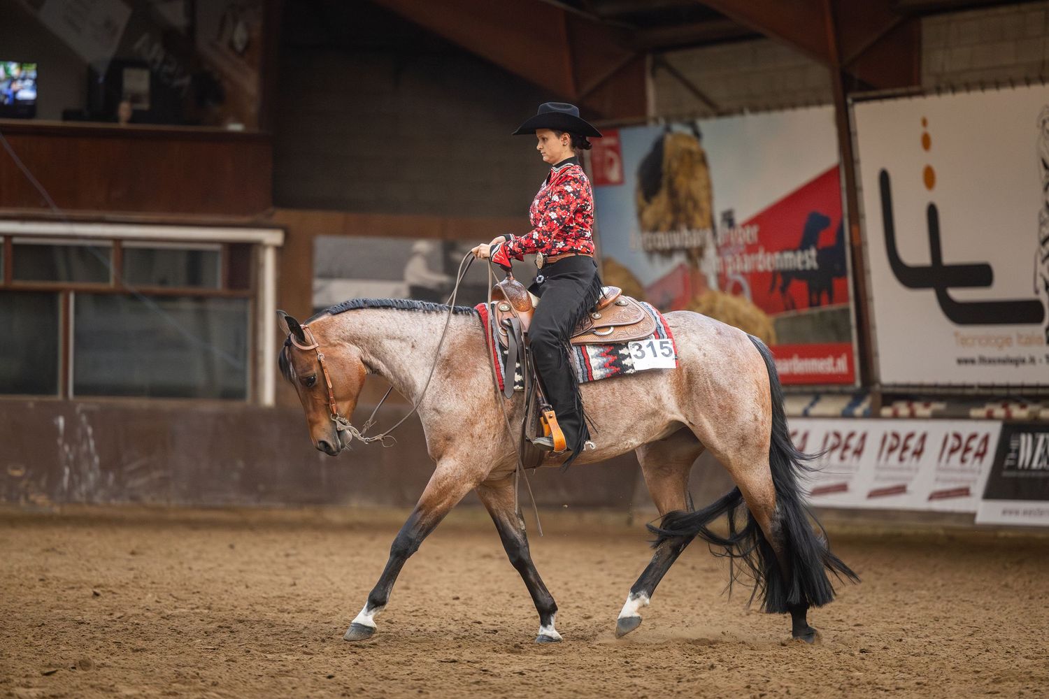 A rider in a red outfit on a palomino horse performing in an indoor western equestrian competition.