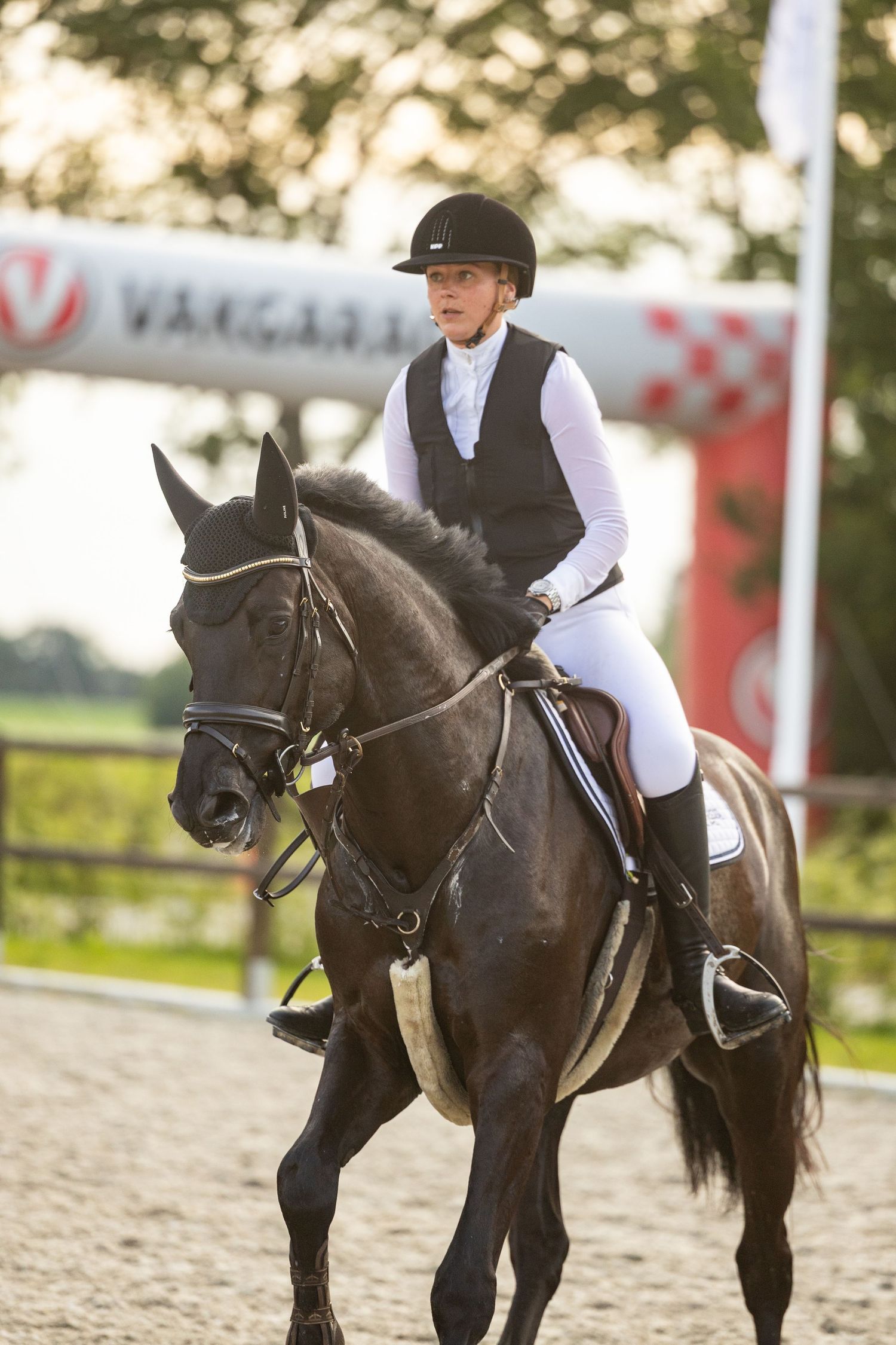Young equestrian rider in black vest and helmet riding a dark horse at an outdoor dressage competition.