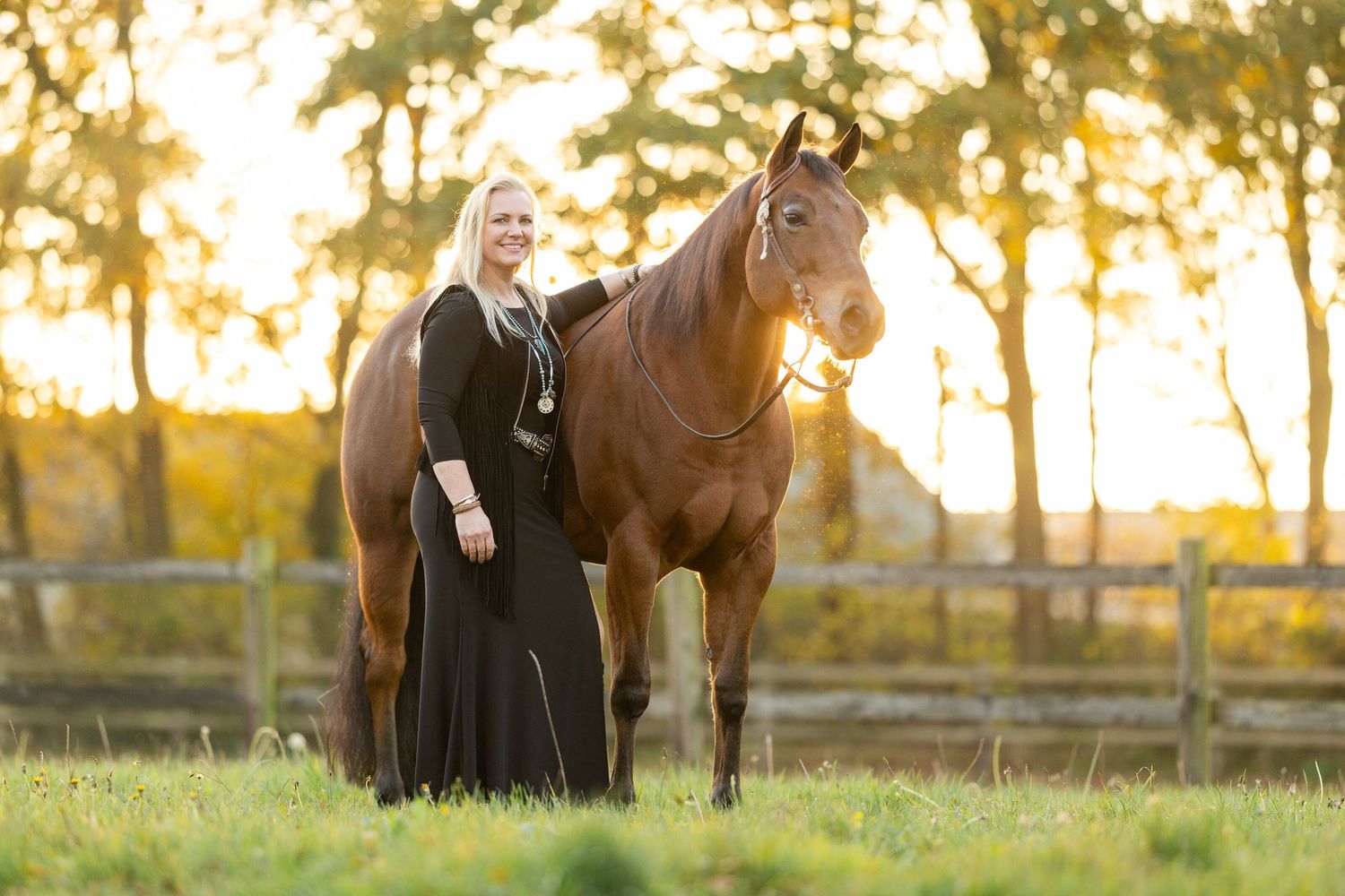 Person in long black dress poses with brown horse during sunset in rural setting.