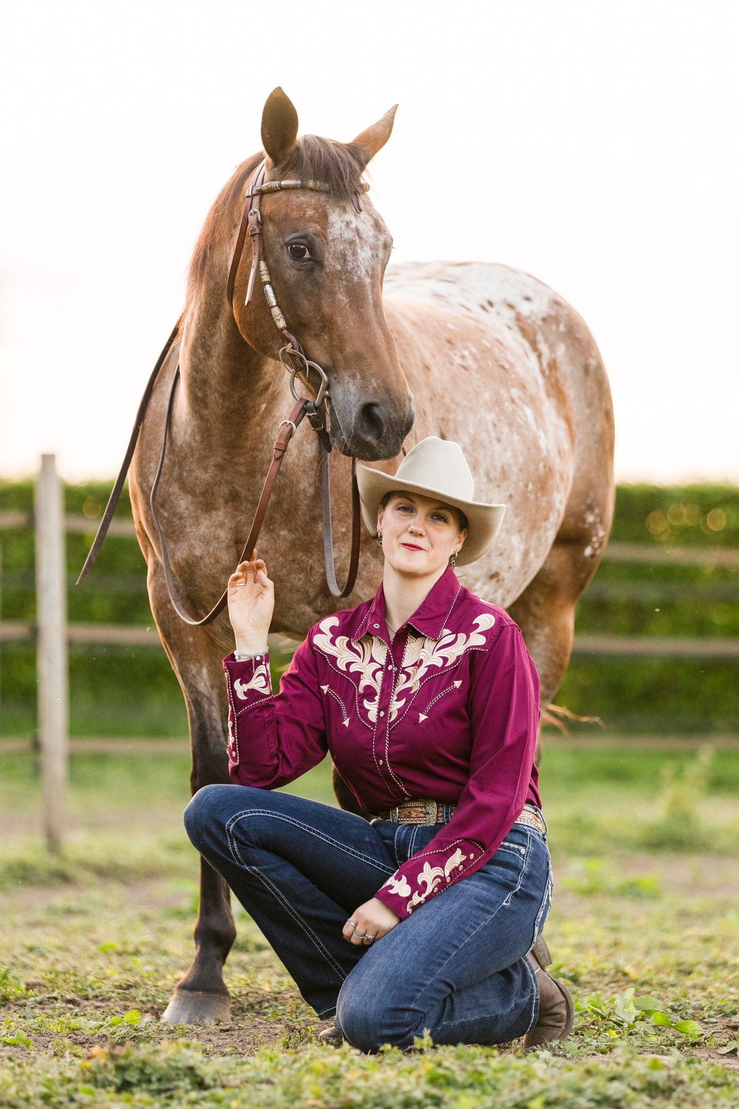 Someone in western wear sits beside a spotted horse in a grassy field at sunset.