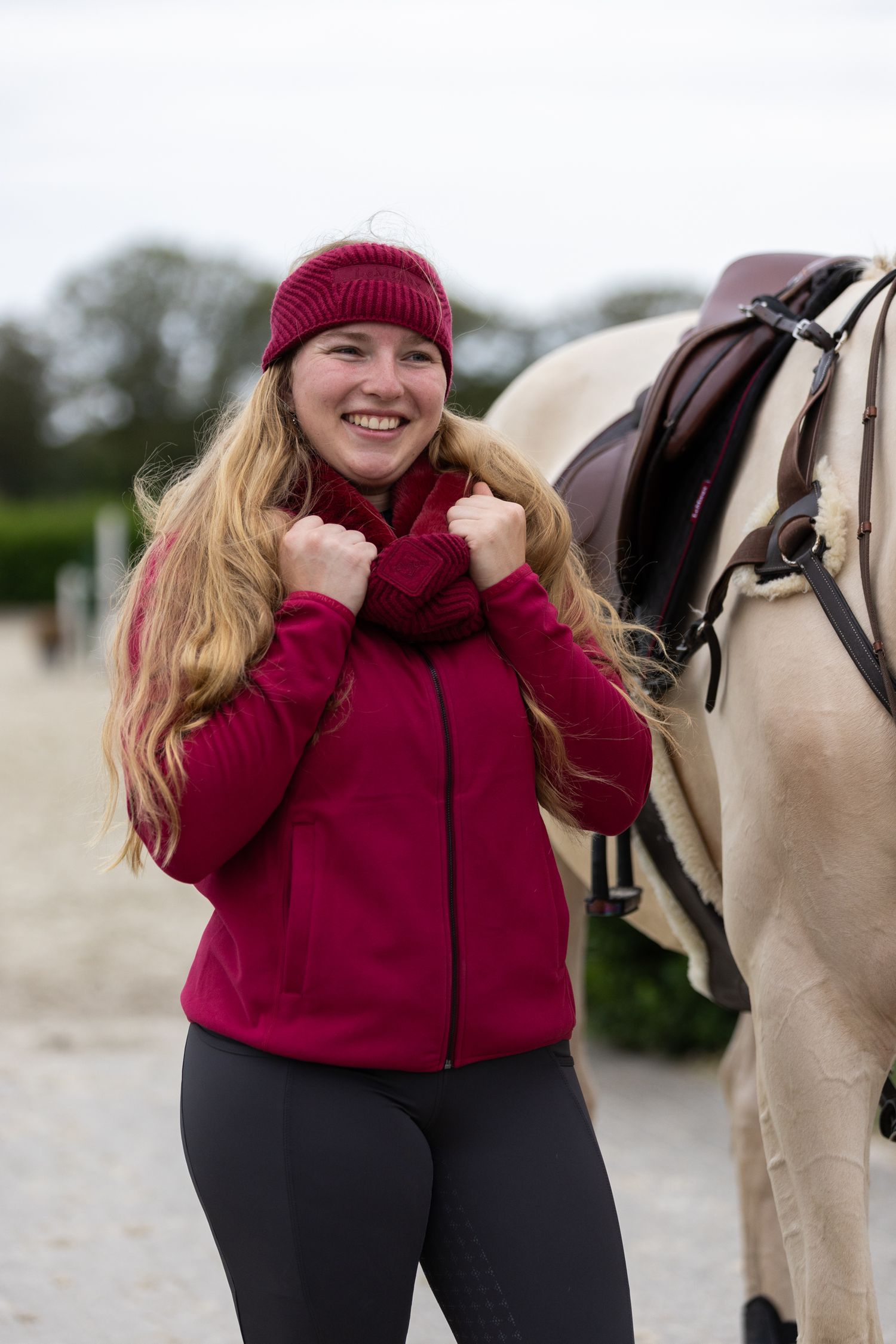 A rider in a burgundy sweater and beanie stands next to a light-colored horse in an outdoor arena.