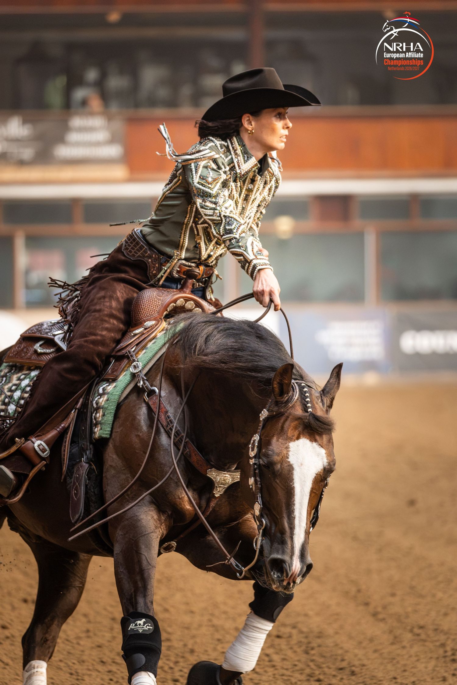 Female rider in decorative western attire competing in NRHA reining event on dark horse in indoor arena.