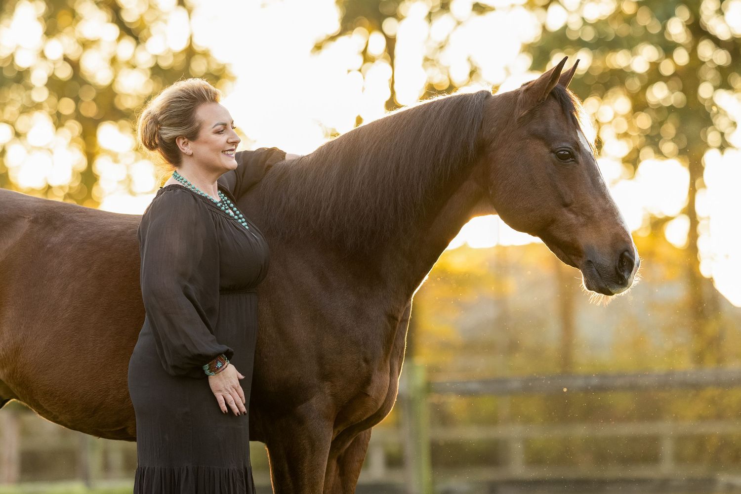 Brown Quarterhorse and person in black dress photographed in warm evening light with trees in background.