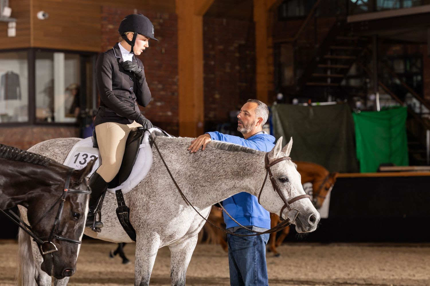 Horses and handlers practicing in an indoor riding arena with wood beam ceiling.