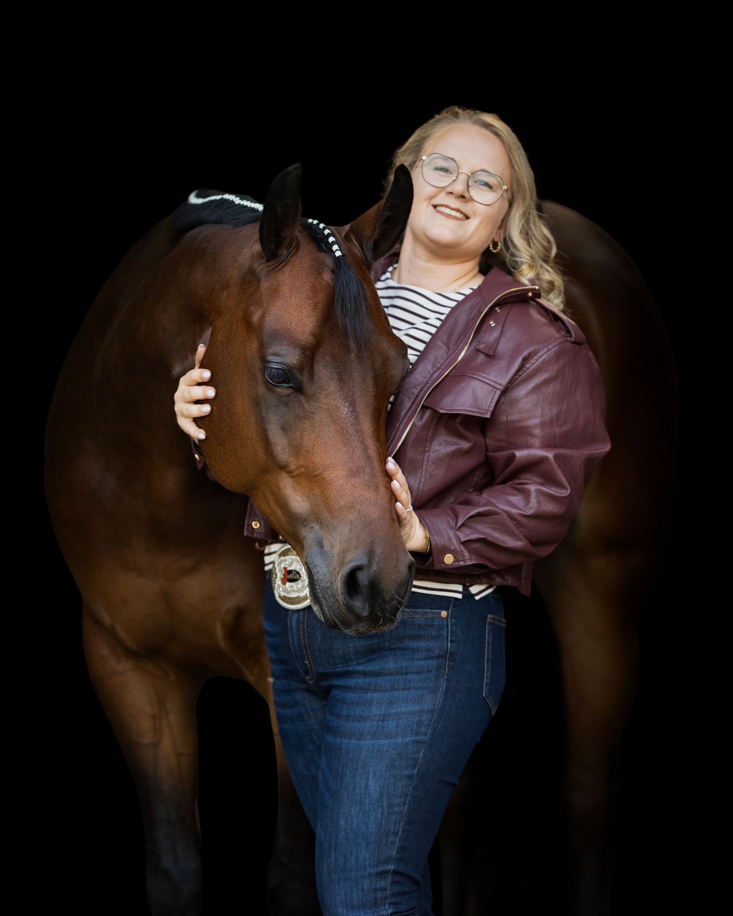 Young woman with glasses in burgundy jacket smiles beside a bay horse against black background.
