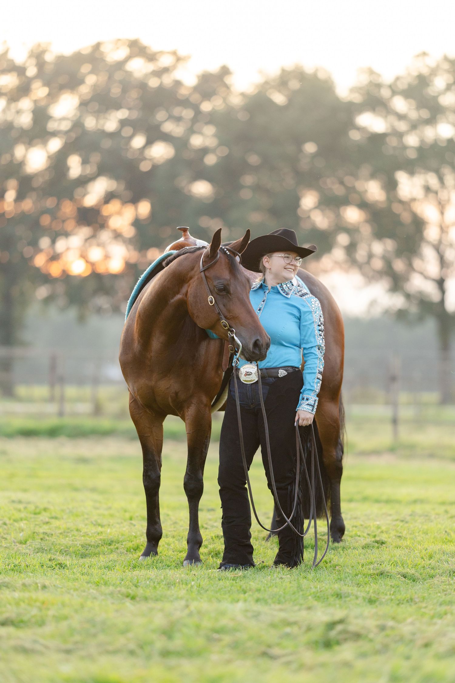 A smiling woman in a blue western shirt and black cowboy hat stands with a chestnut horse on a green field at sunset.