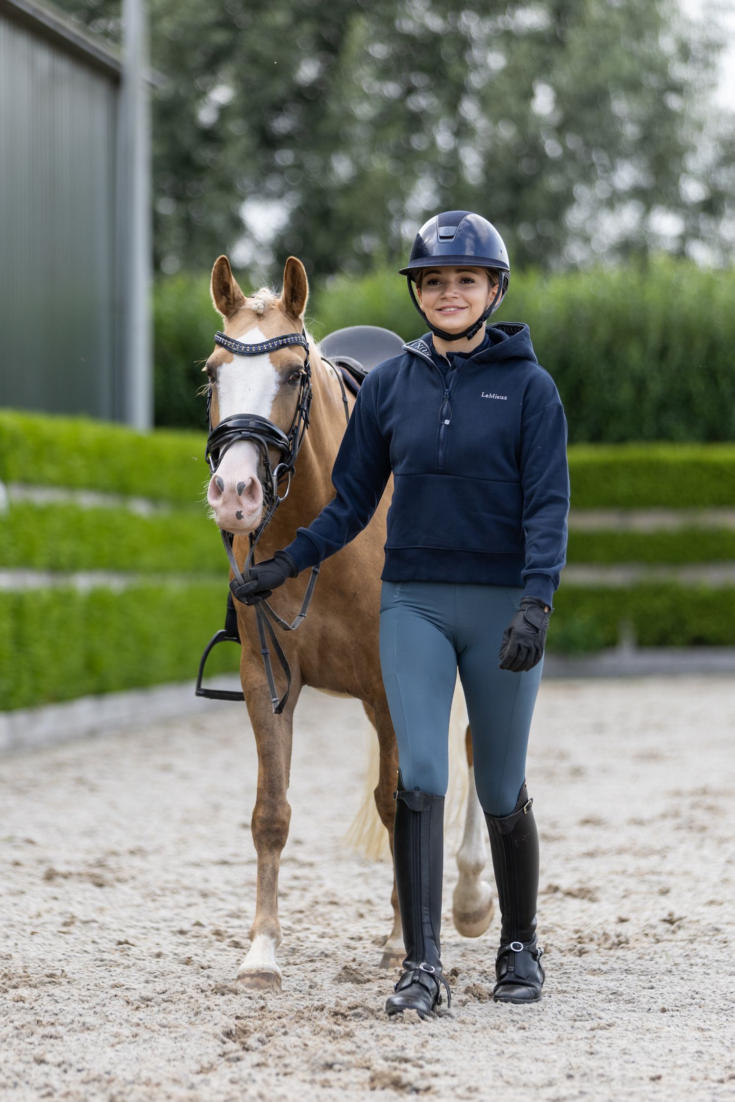 Brown and white pony with handler standing in a sandy arena wearing riding gear.