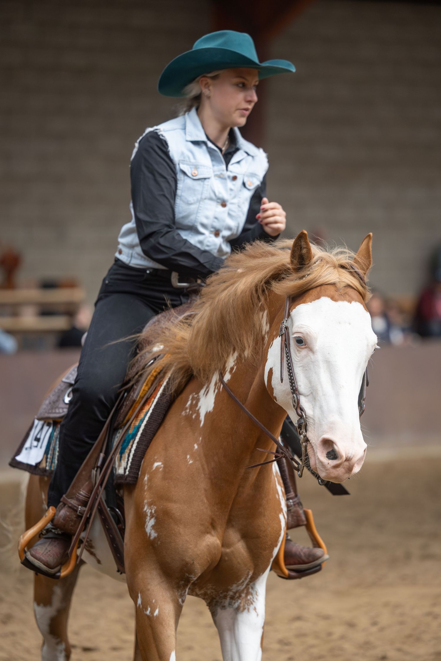 Young woman in teal cowboy hat riding a chestnut and white paint horse in an indoor western riding arena.