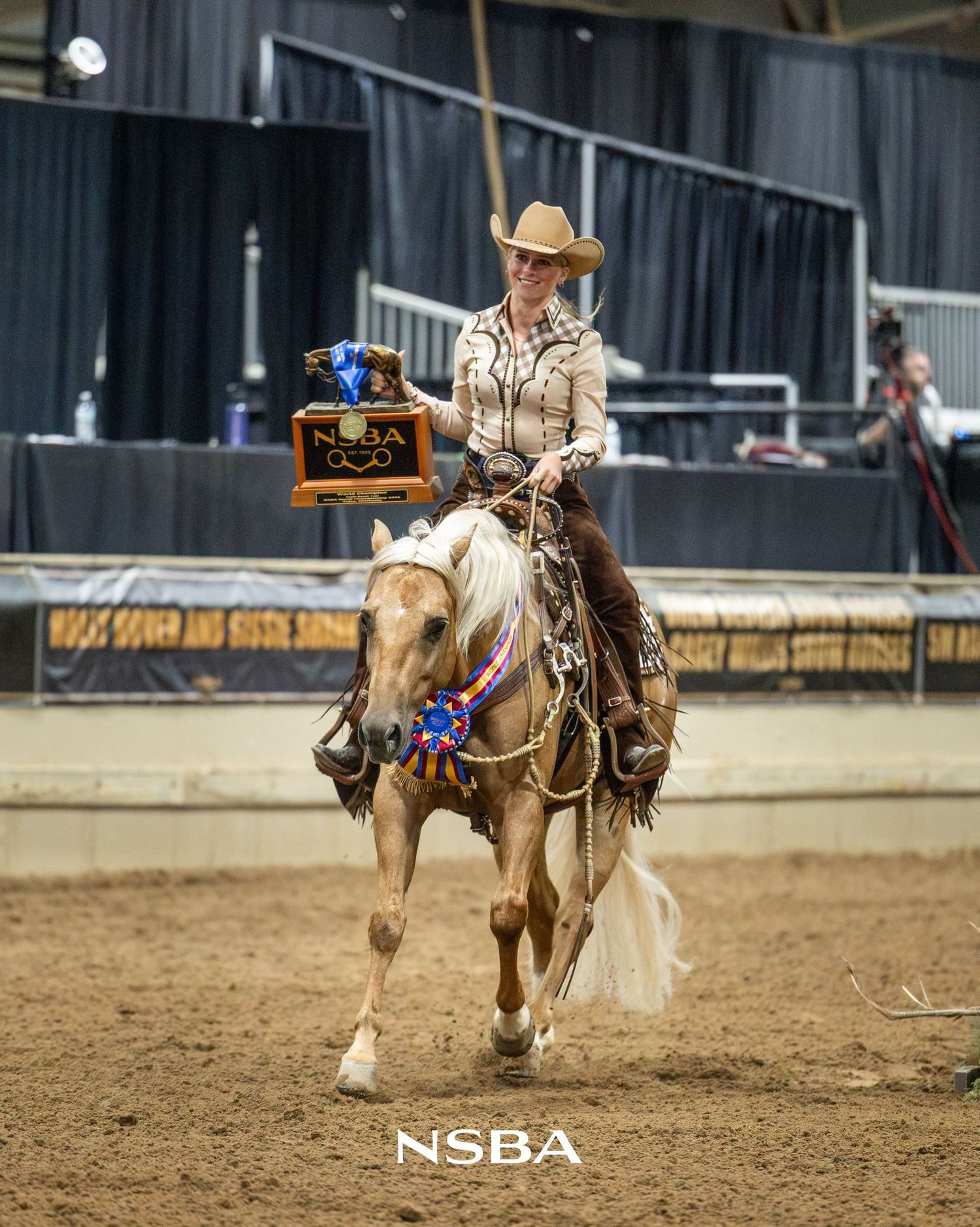 Woman in cowboy hat riding palomino horse holding NSBA trophy at indoor equestrian competition.