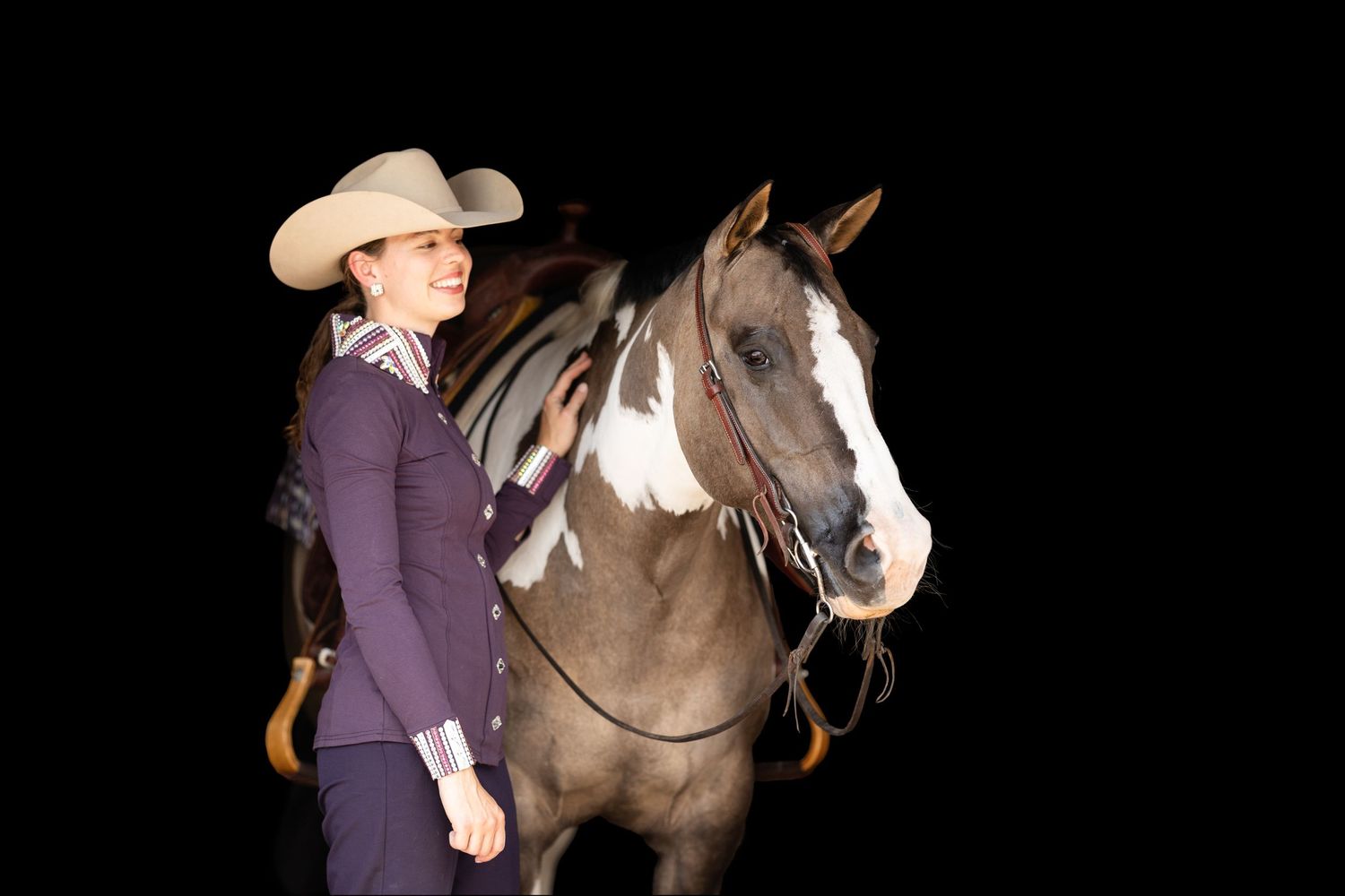 A paint horse stands with a rider wearing a purple show outfit against a black background.