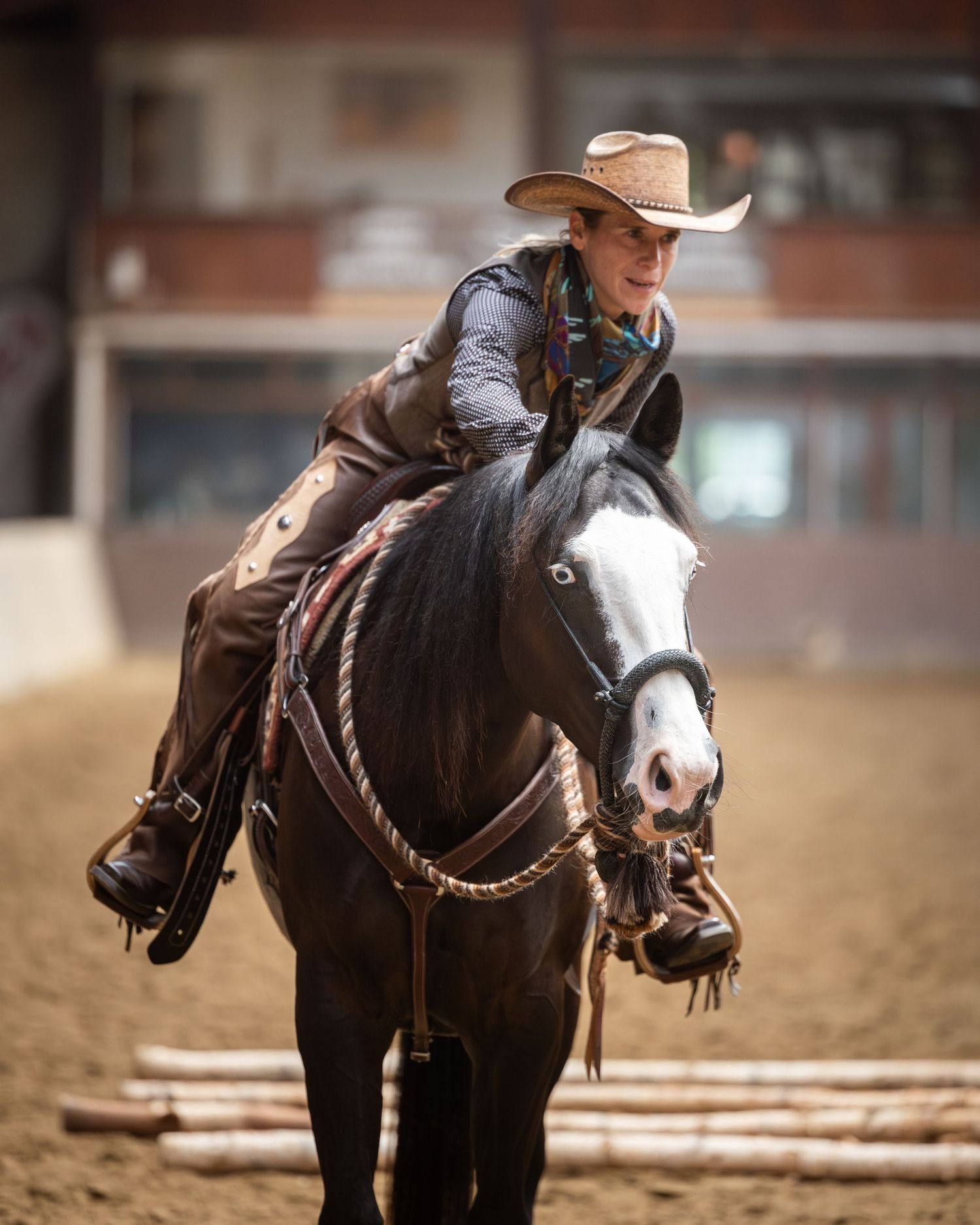 Cowboy hat-wearing rider on a dark horse competing in an indoor western equestrian event on sandy arena ground.