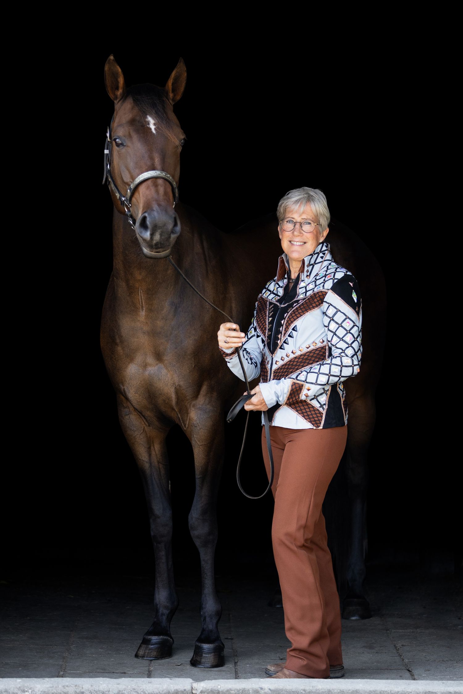 Person in brown pants and patterned shirt posing with a brown horse in a dark studio setting.