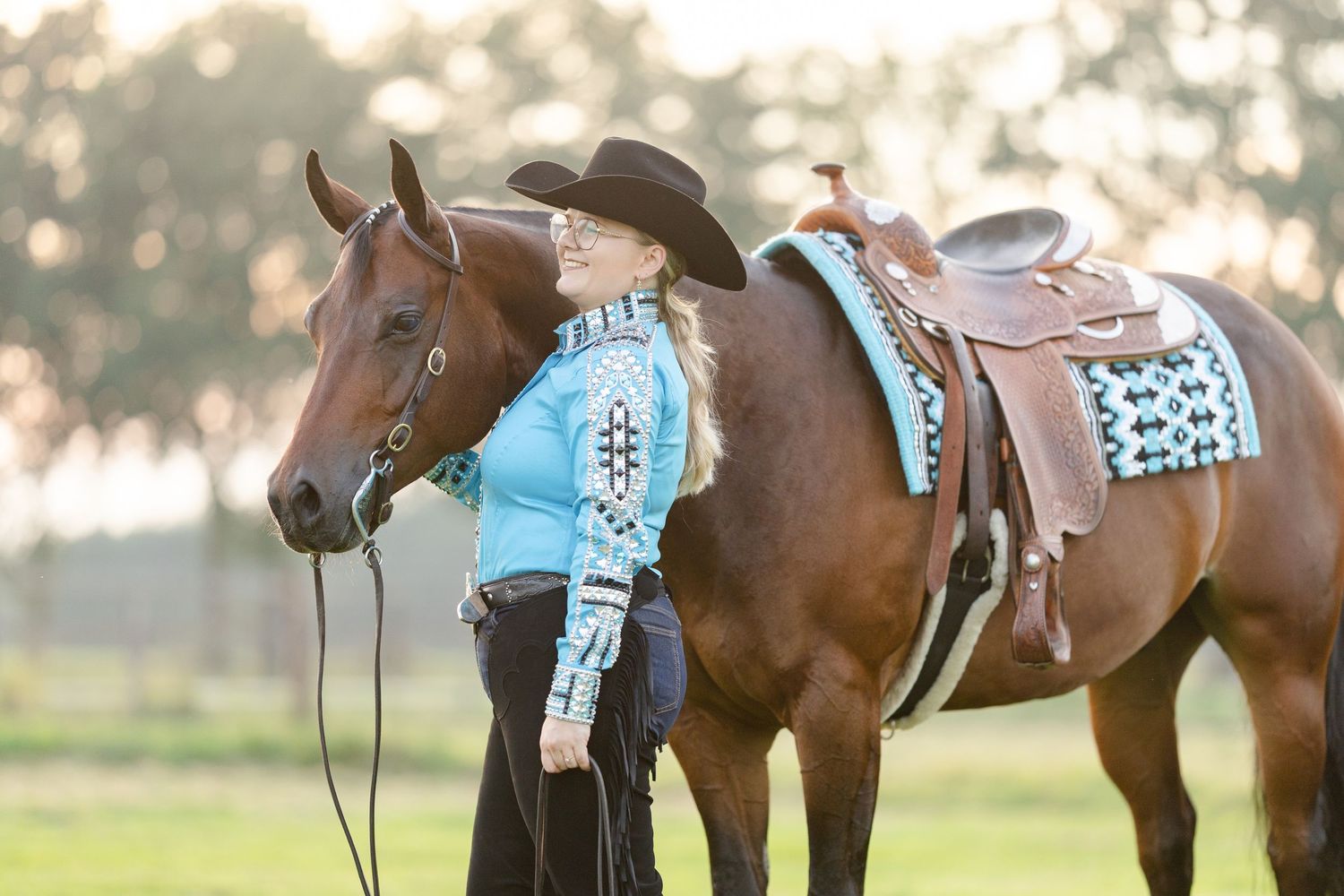 Blonde woman in turquoise western shirt and black cowboy hat standing beside a chestnut horse with teal saddle pad.
