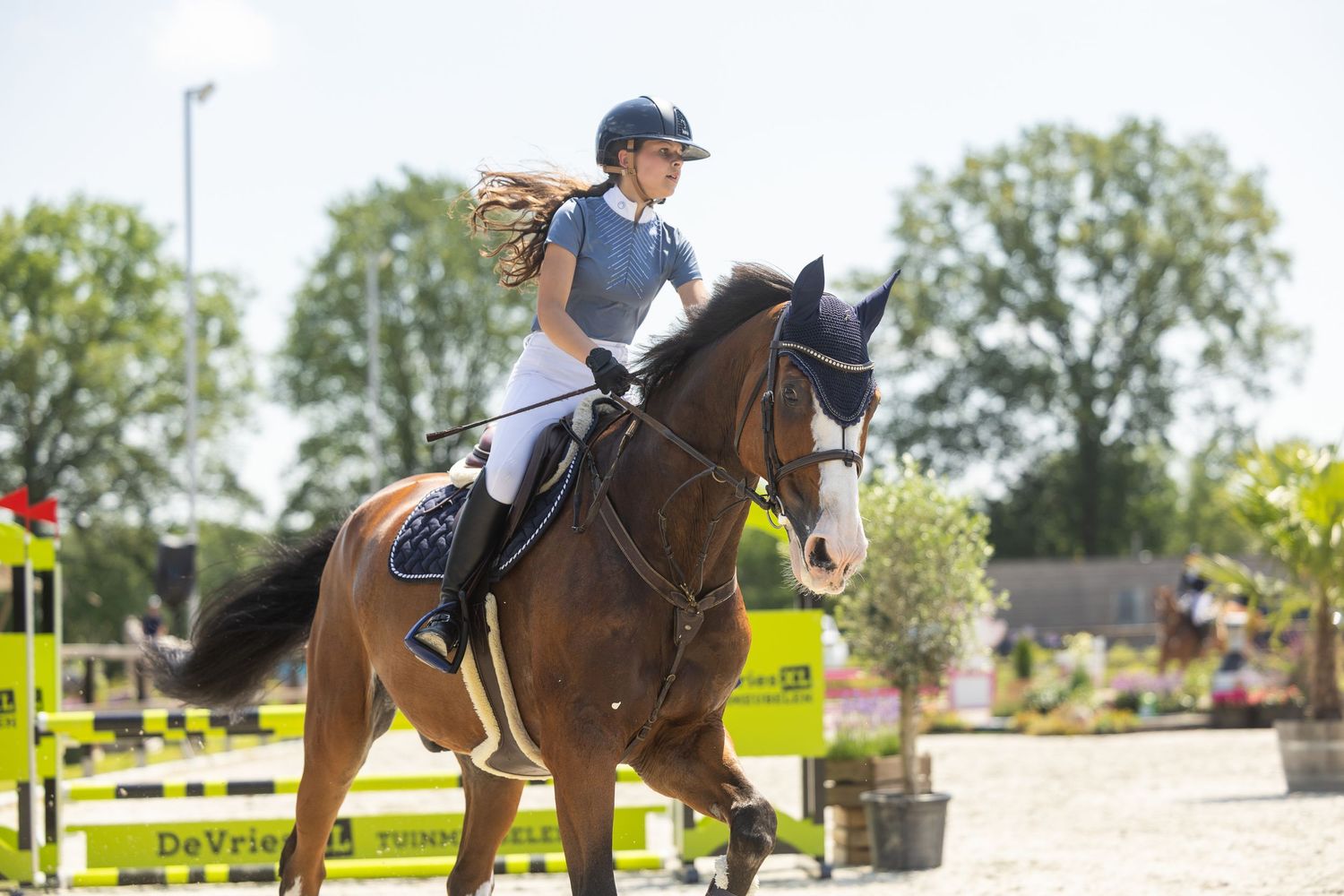Young female equestrian in helmet rides chestnut horse at outdoor show jumping competition on sunny day.