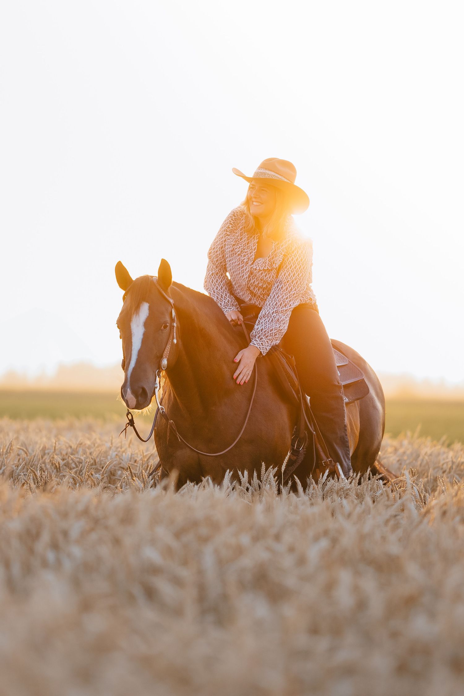 A cowgirl in a hat rides a chestnut horse through a golden wheat field at sunset.