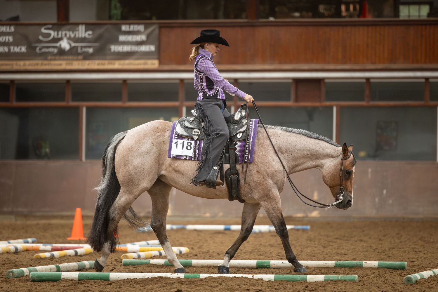 Rider in purple shirt on gray horse competing in western equestrian event inside indoor arena with number 118.