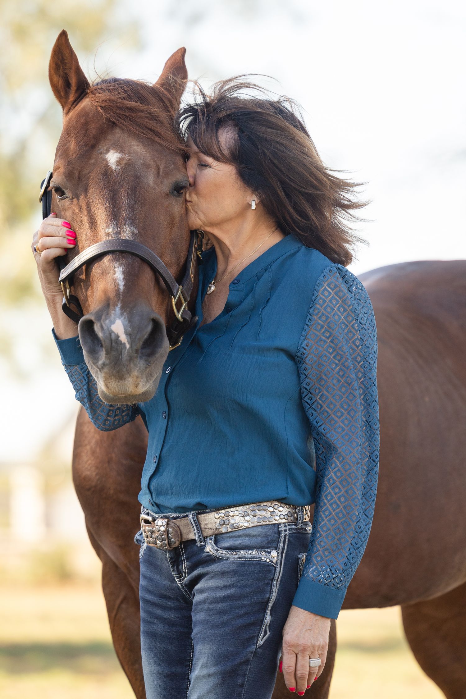 Person in blue sweater giving affectionate kiss to brown horse outdoors.