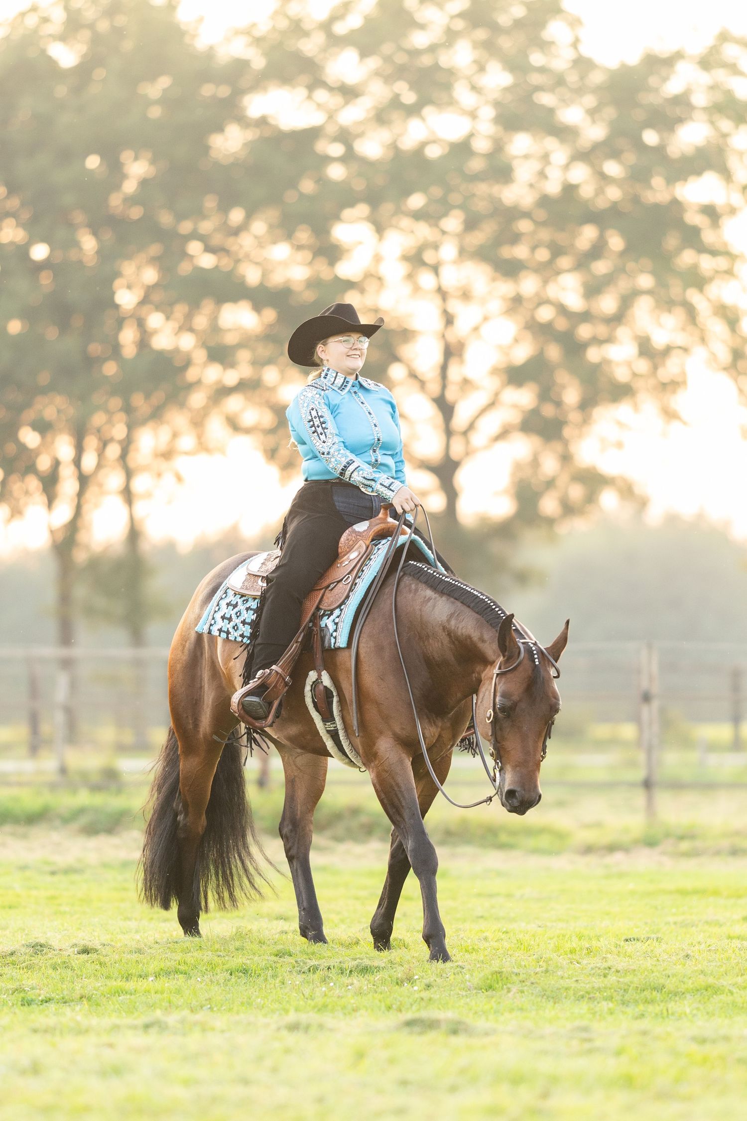Woman in blue western shirt and cowboy hat riding a chestnut horse on green grass at golden hour.