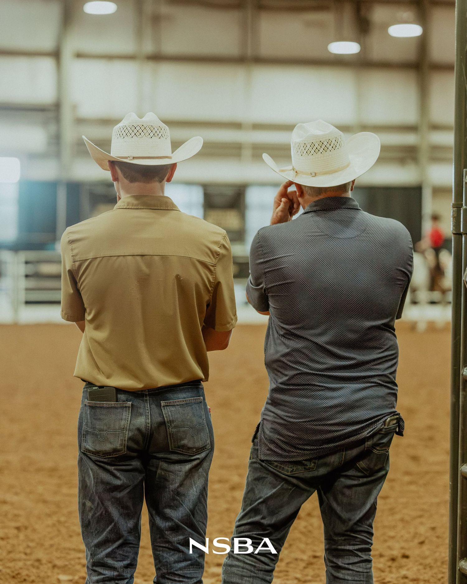 Two cowboys in white straw hats and jeans stand with backs turned at an NSBA equestrian arena event.
