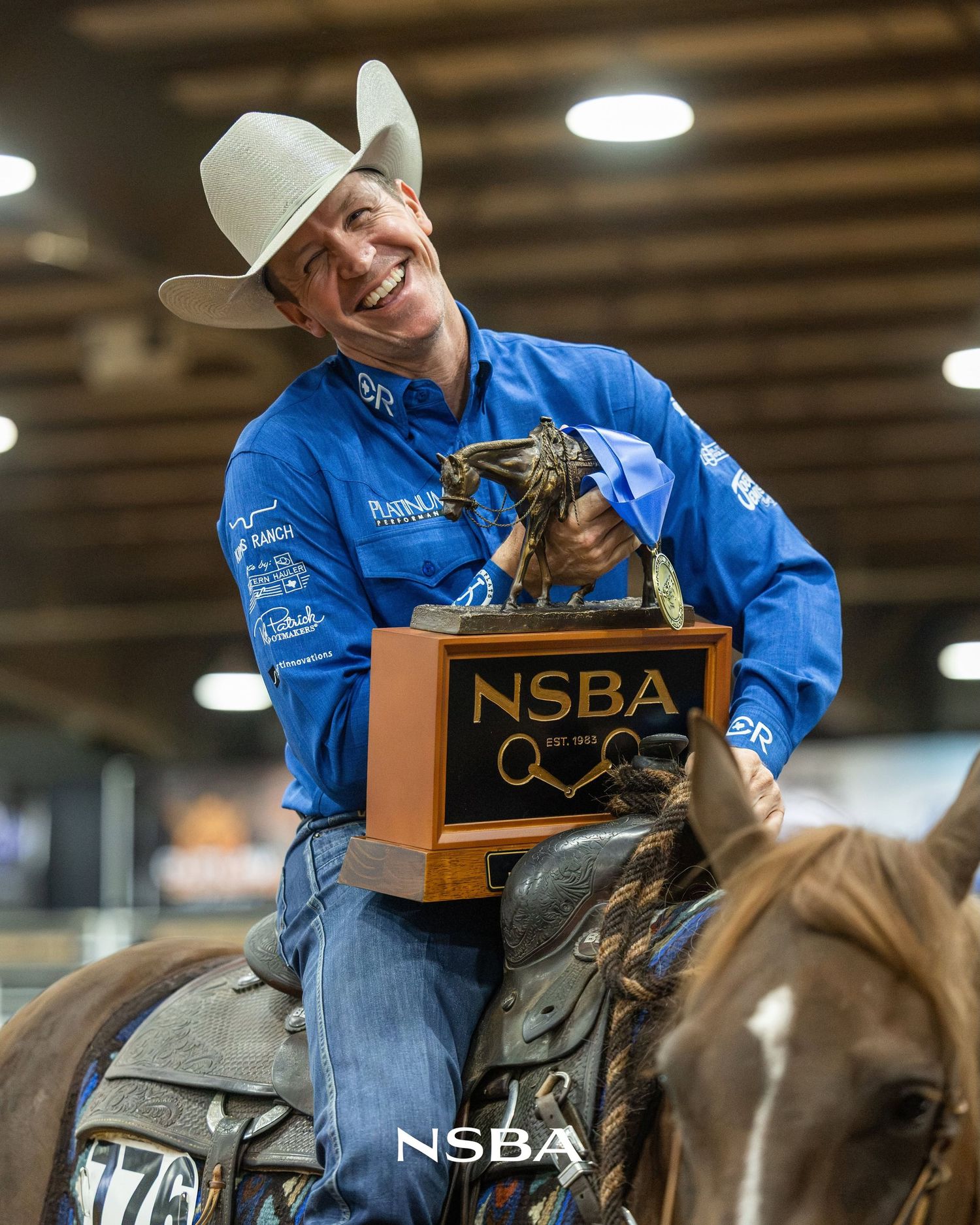 Smiling Bud Lyon in blue jacket and white hat holds NSBA trophy while seated on horseback at an equestrian event.
